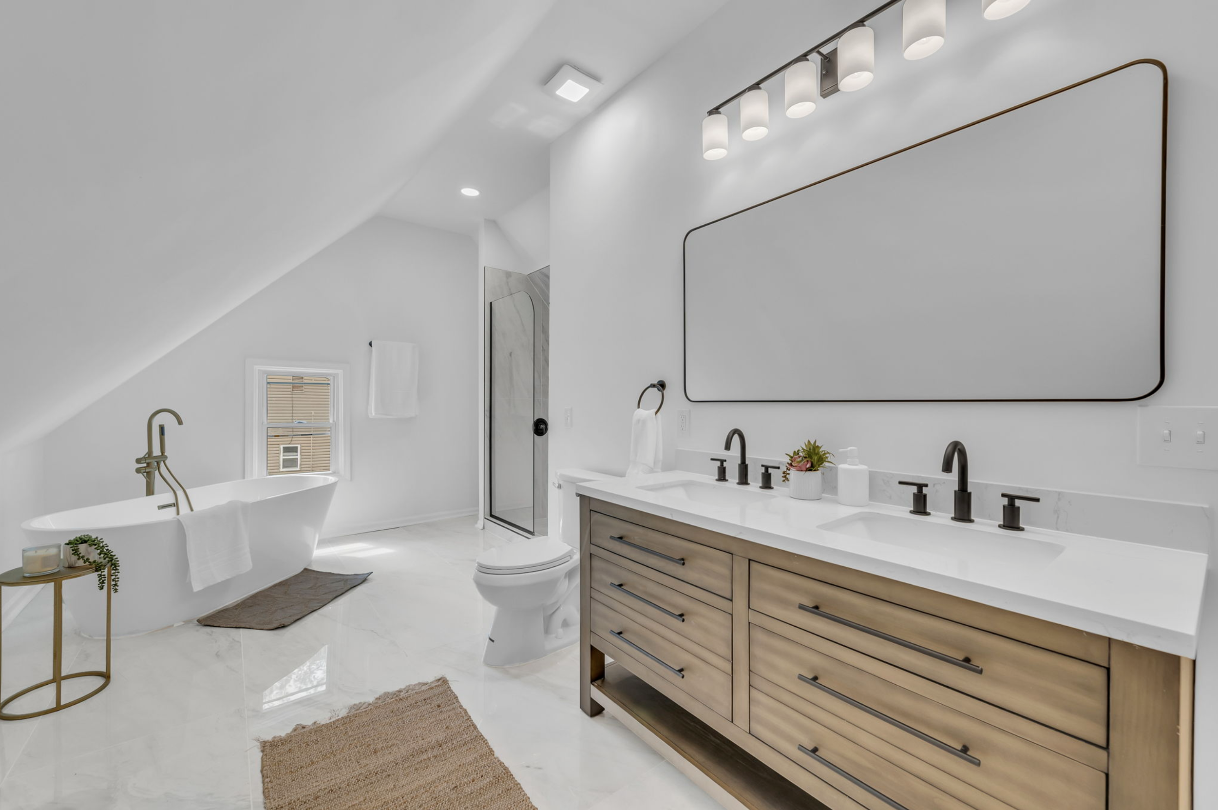 Modern white bathroom with a freestanding bathtub, a double vanity with a large mirror, a glass-enclosed shower, and neutral towels and rugs.
