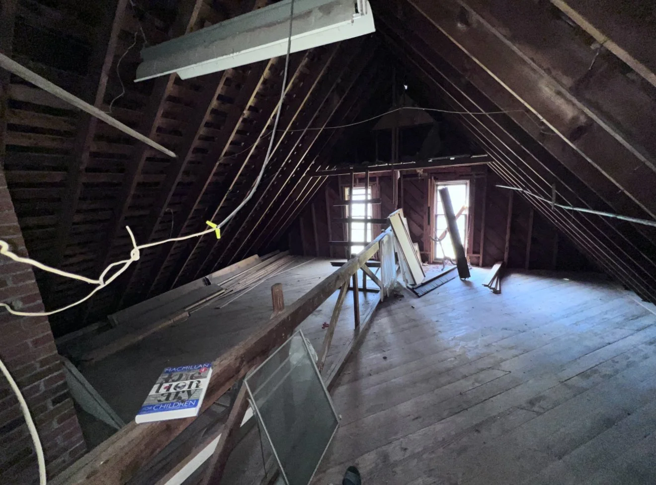 Empty attic with slanted wooden ceiling, scattered construction items, ladder, and windows at the far end, with natural light coming through.