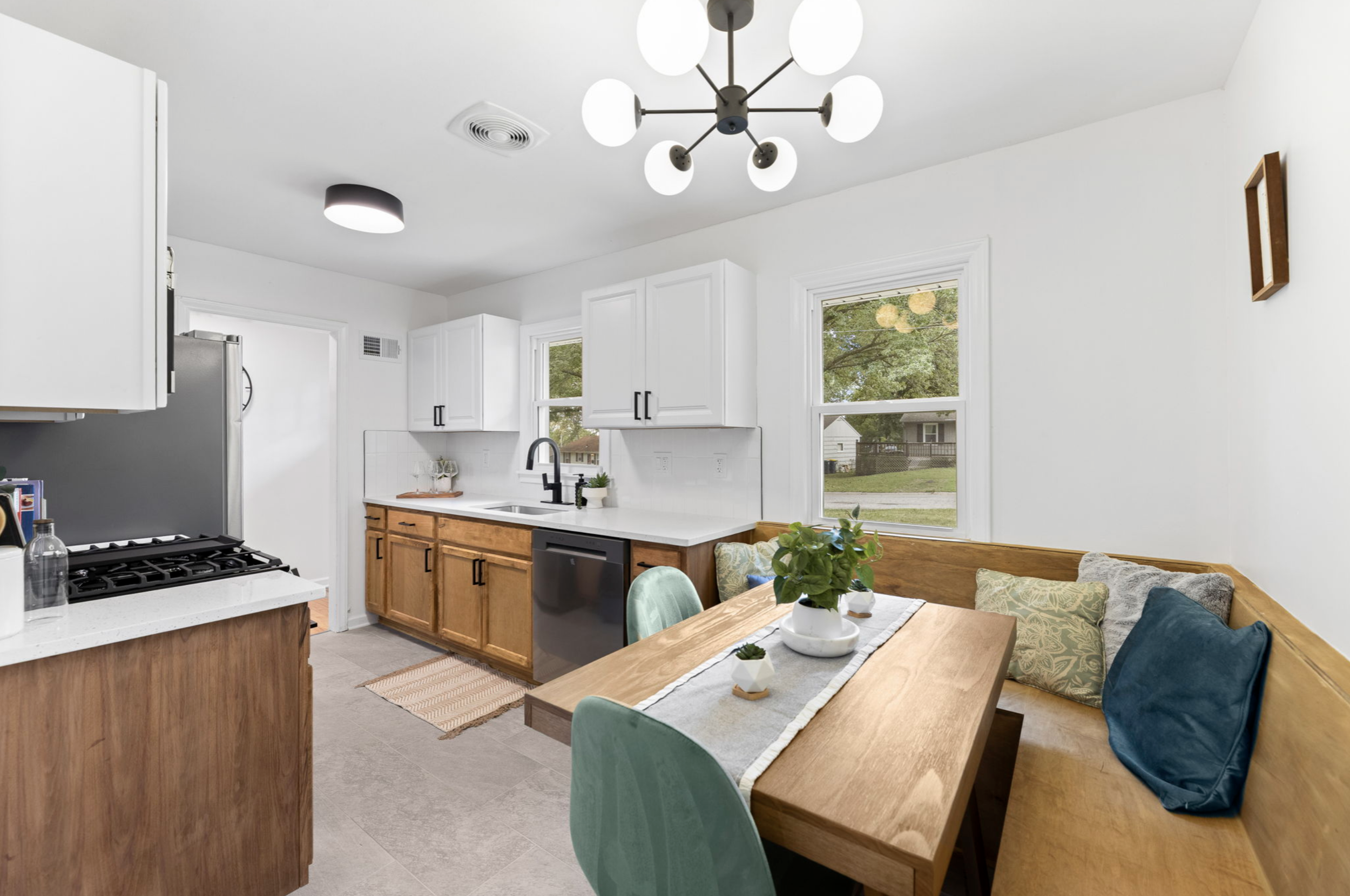 Modern kitchen with white cabinets, wooden lower cabinets, black fixtures, and a dining nook with a wooden table, green and blue chairs, and a cushioned bench near two windows