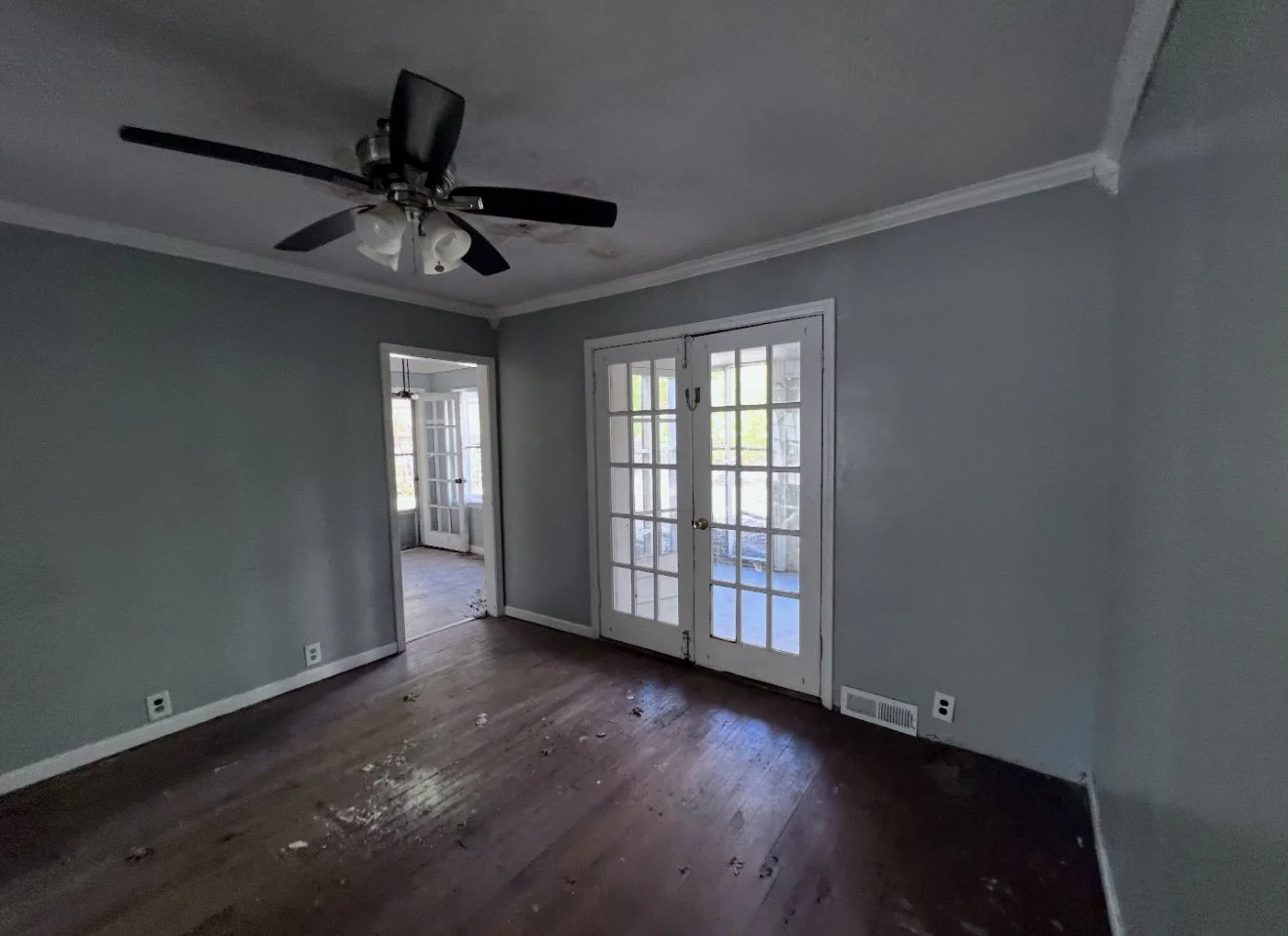 Empty living room with wood flooring, gray walls, a ceiling fan, French doors leading outside, and a doorway to another room with a view of a porch.