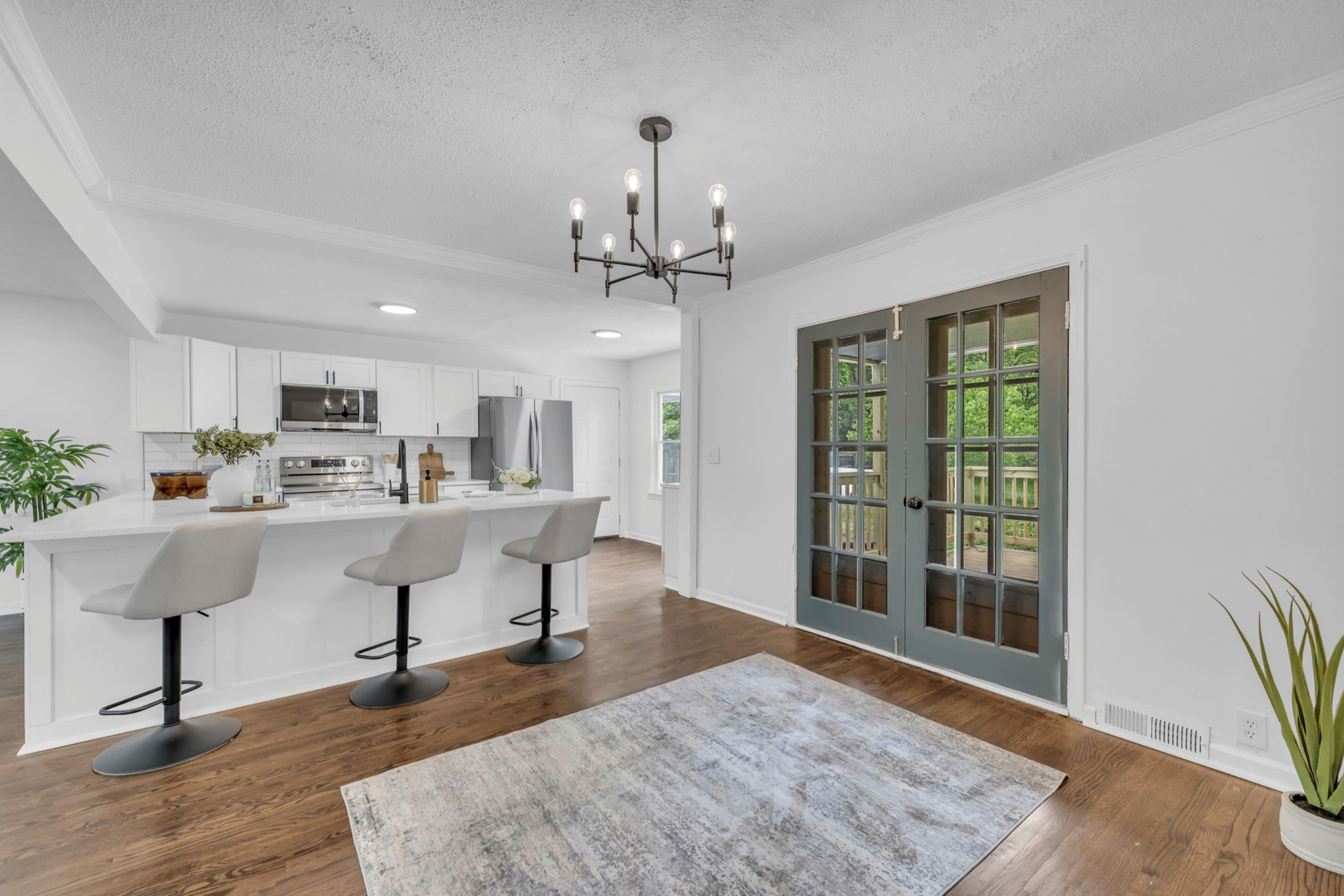 Living area with French doors leading to an outdoor space, hardwood floors, a patterned area rug, a hanging black chandelier, and a kitchen with white cabinets, stainless steel appliances, and a white island with three beige bar stools.