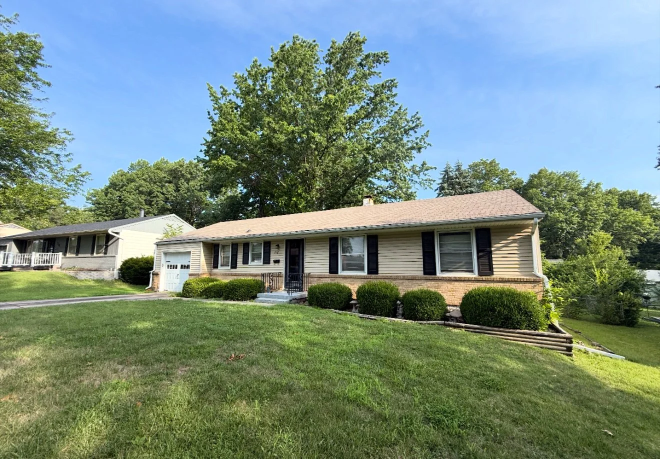 Single-story house with beige siding, black shutters, and a garage, surrounded by a green lawn and bushes, with large trees in the background under a blue sky.