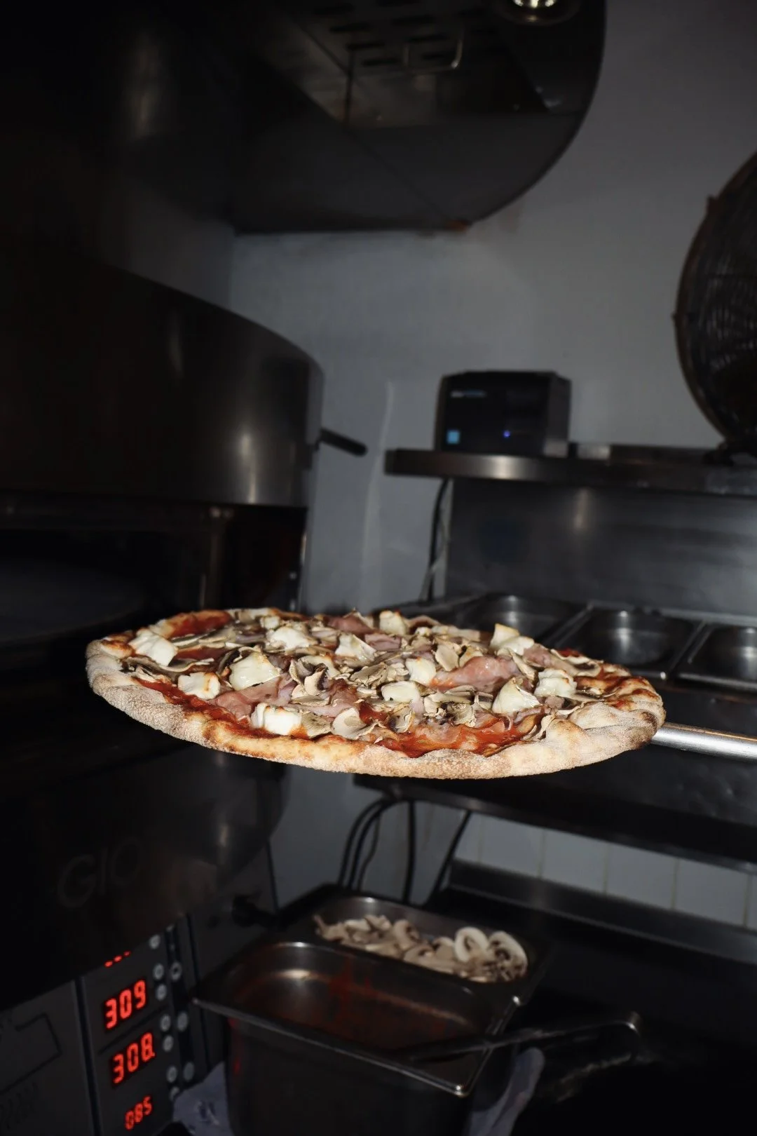 Een pizza met champignons, ham en kaas wordt uit de oven gehaald in een professionele keuken.