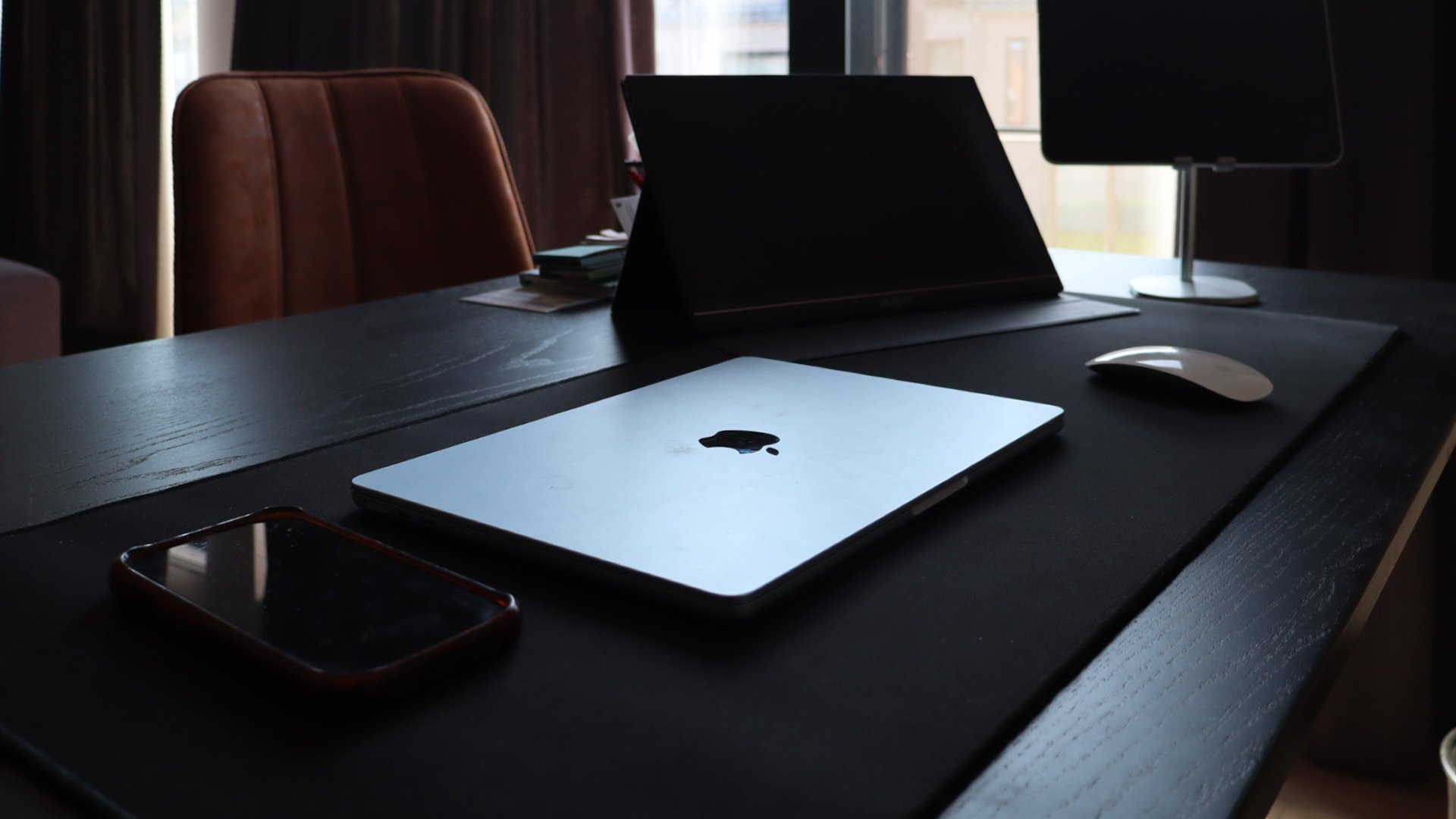 Een donkere tafel met een open MacBook, een witte muis, een smartphone, een laptop met het Apple logo, een bureaulamp en enkele papieren op de achtergrond.