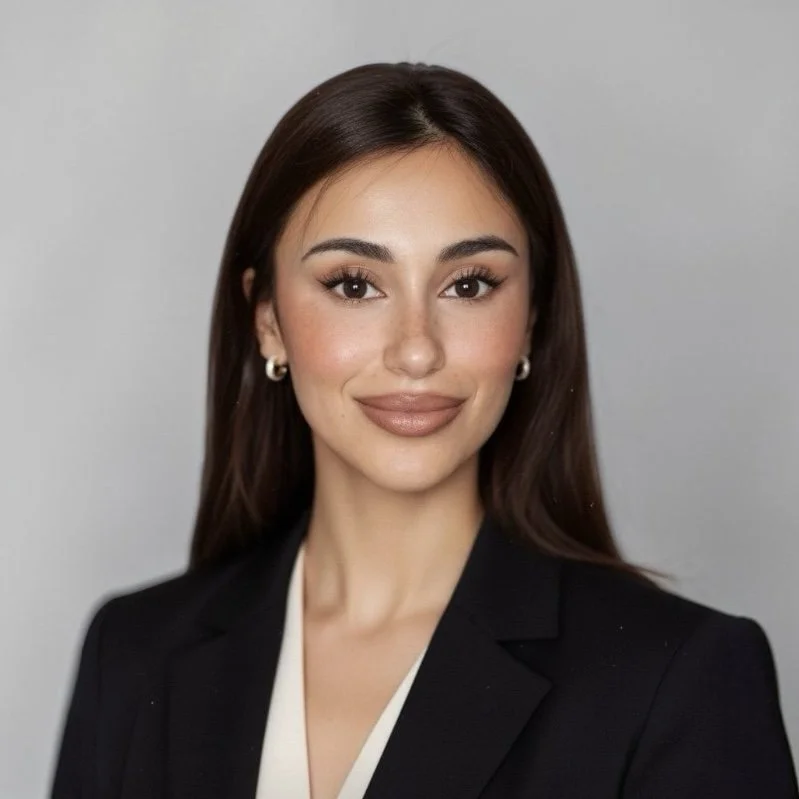 Portret van een jonge vrouw met lang bruin haar, smokey eye make-up, lichtbruine lipstick, in een zwarte blazer en witte blouse, lachend voor een grijze achtergrond.