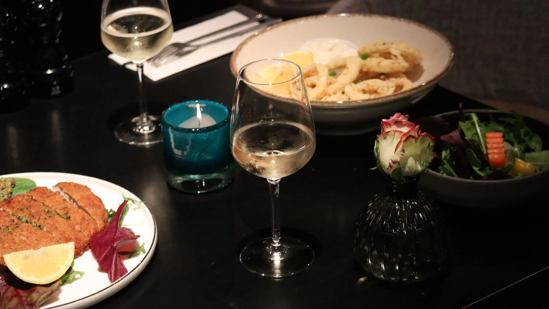 Een black table met een glas witte wijn, een glas champagne, een bord met een gepaneerd gerecht met citroen en salade, een kom met risotto of pasta, en een kleine vaas met een roos en een salade. Er is ook een blauwe kaars en een wit servet.