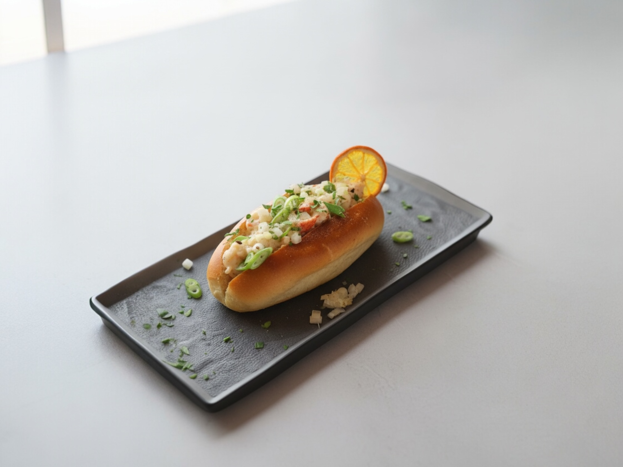 Scampi roll in a soft bun with chopped onions, green onions, and a slice of orange on a black rectangular plate