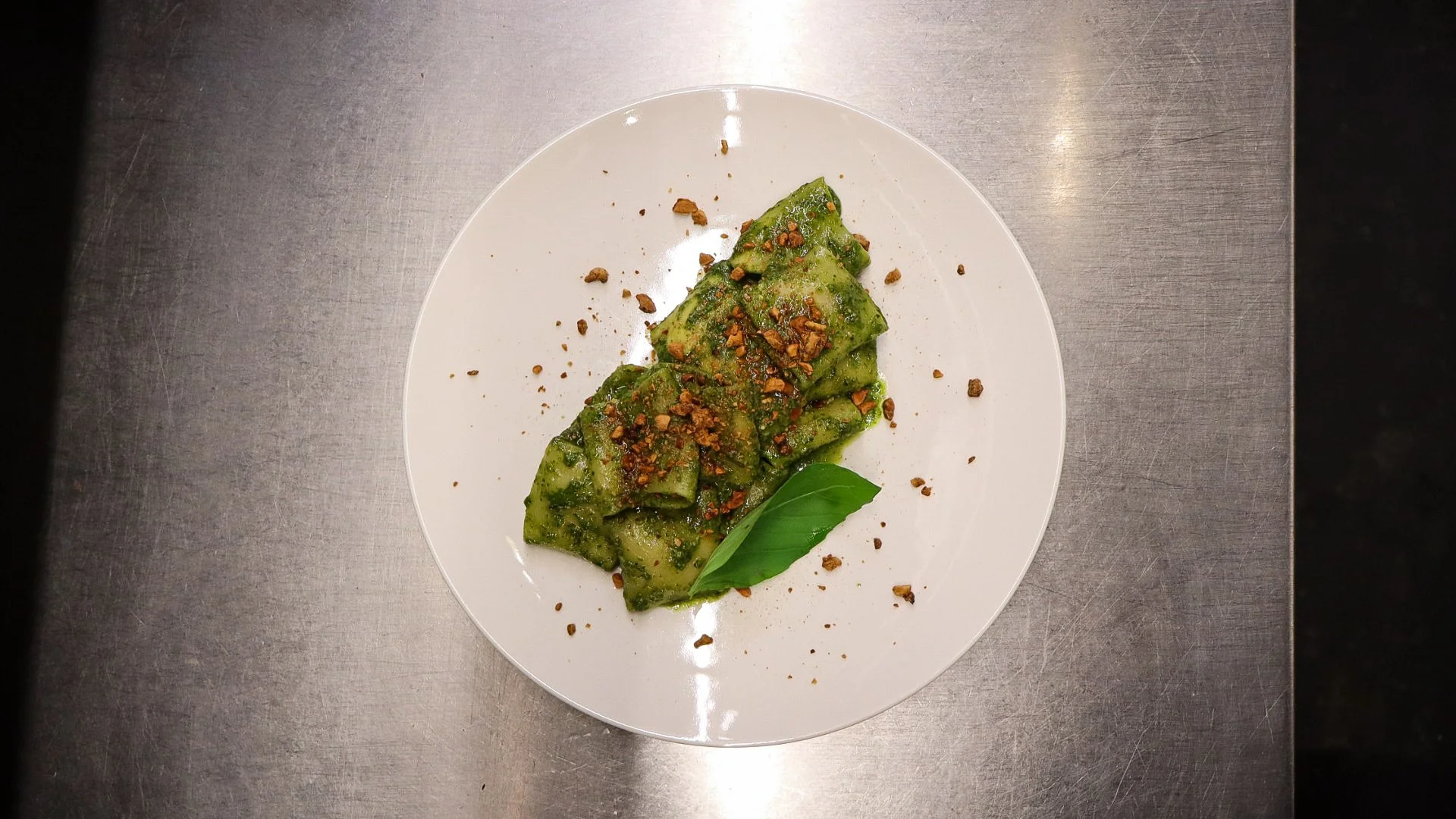 Groene pasta met kruiden en geschaafde noten, versierzeld met een basilicablad, op een wit bord op een metalen oppervlak.