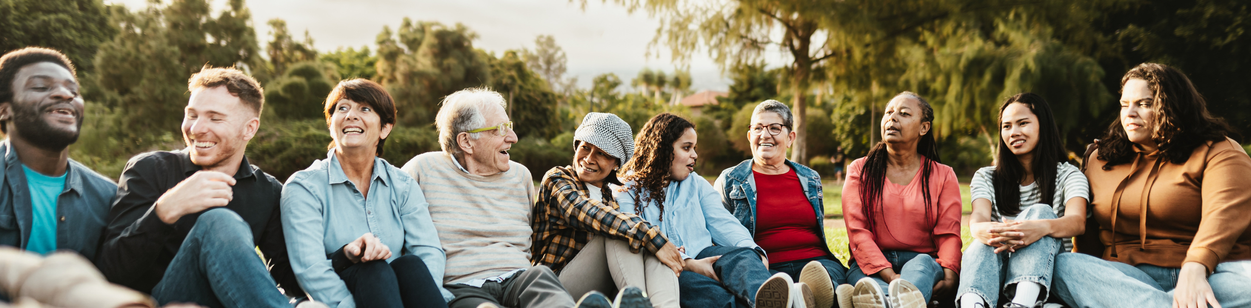 A diverse group of people sitting outside on the grass, engaging and smiling with each other during the daytime.