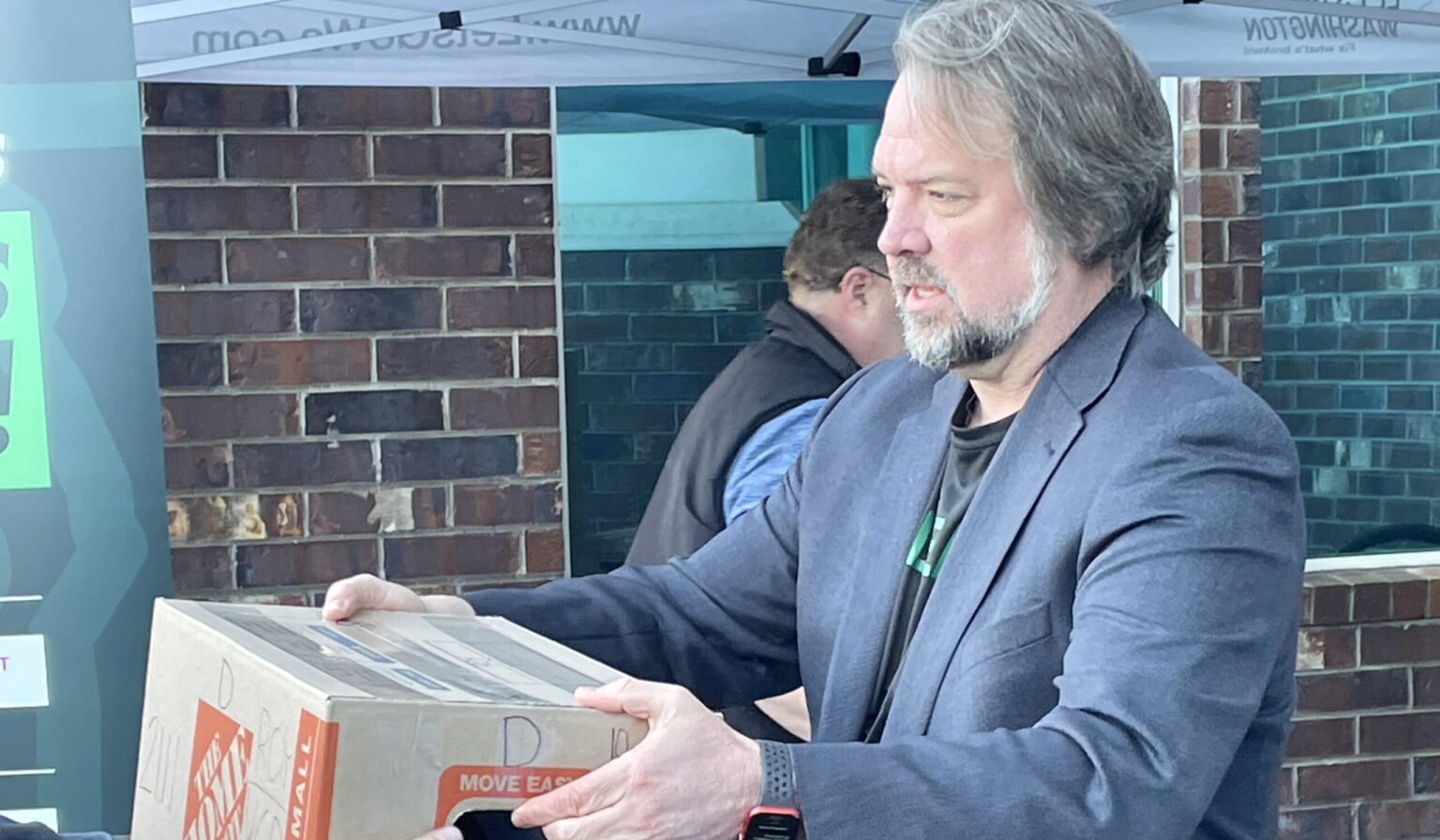 A man with gray hair and a beard wearing a dark gray blazer holding a box with an orange label in front of a brick wall outside.