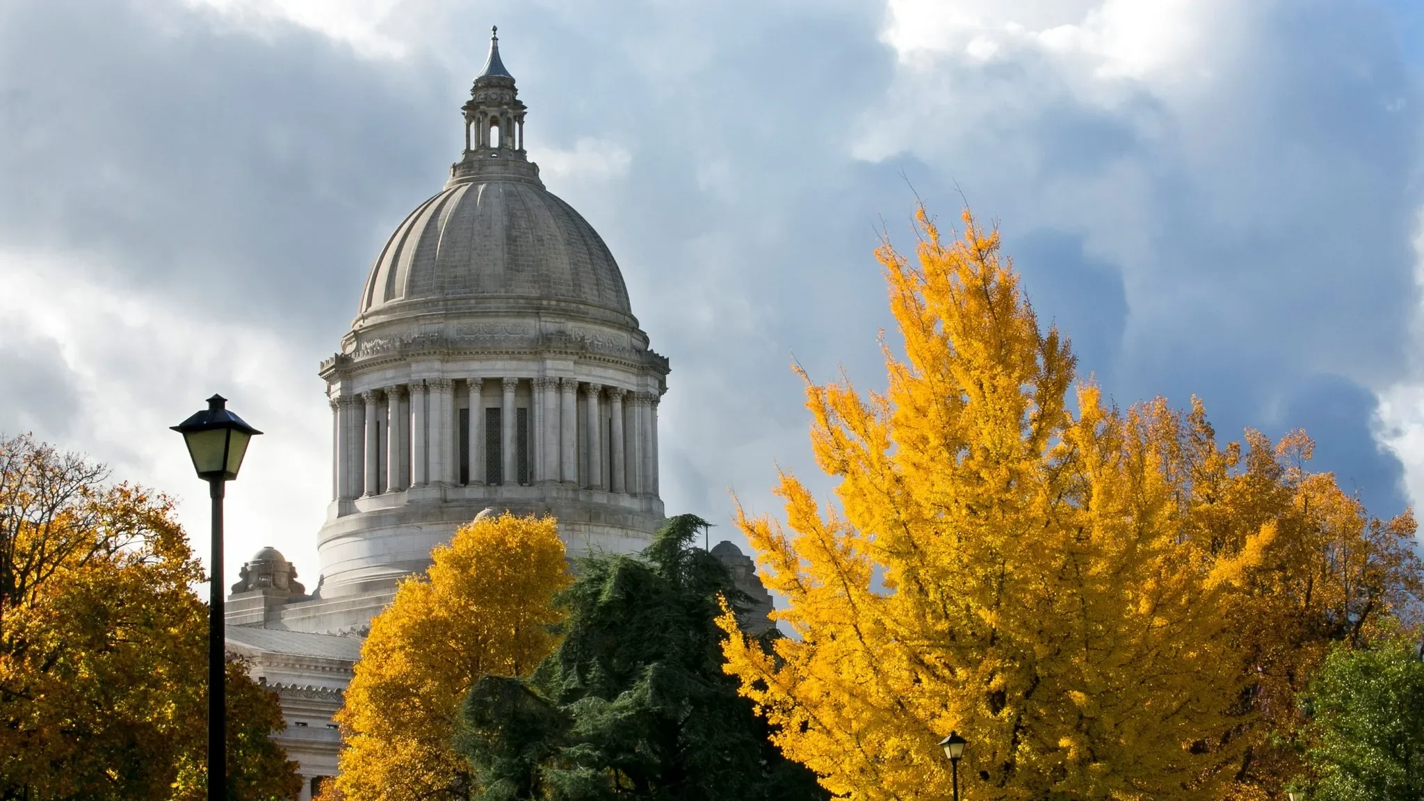 A large domed government building, possibly a capitol, is surrounded by colorful autumn trees with vibrant yellow and orange leaves, and a street lamp in the foreground, under a partly cloudy sky.