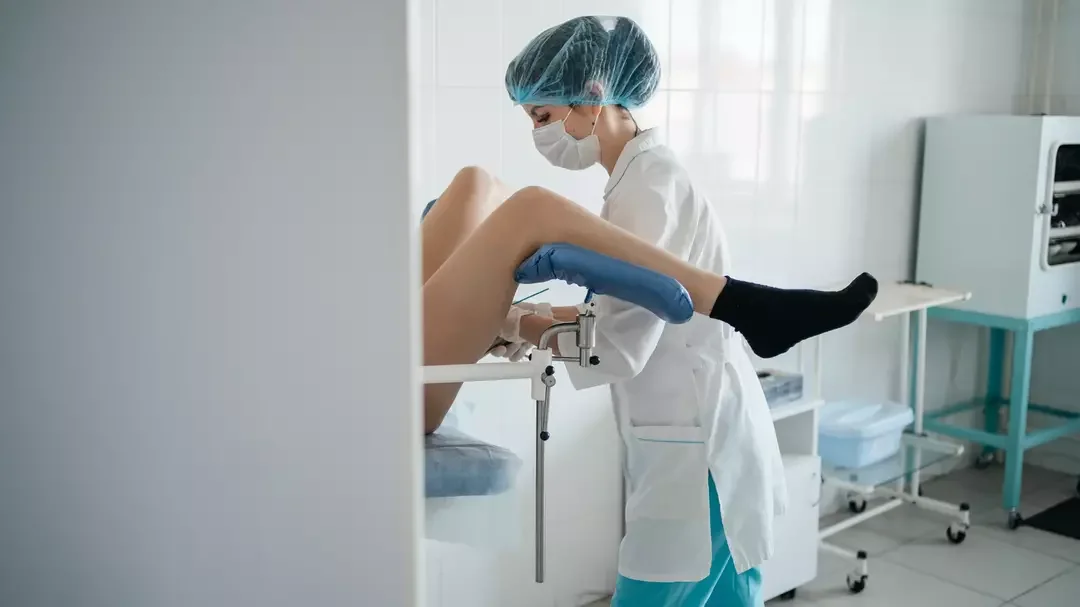 A healthcare worker, wearing a mask, gloves, and a hair cover, assisting a patient in a medical setting.