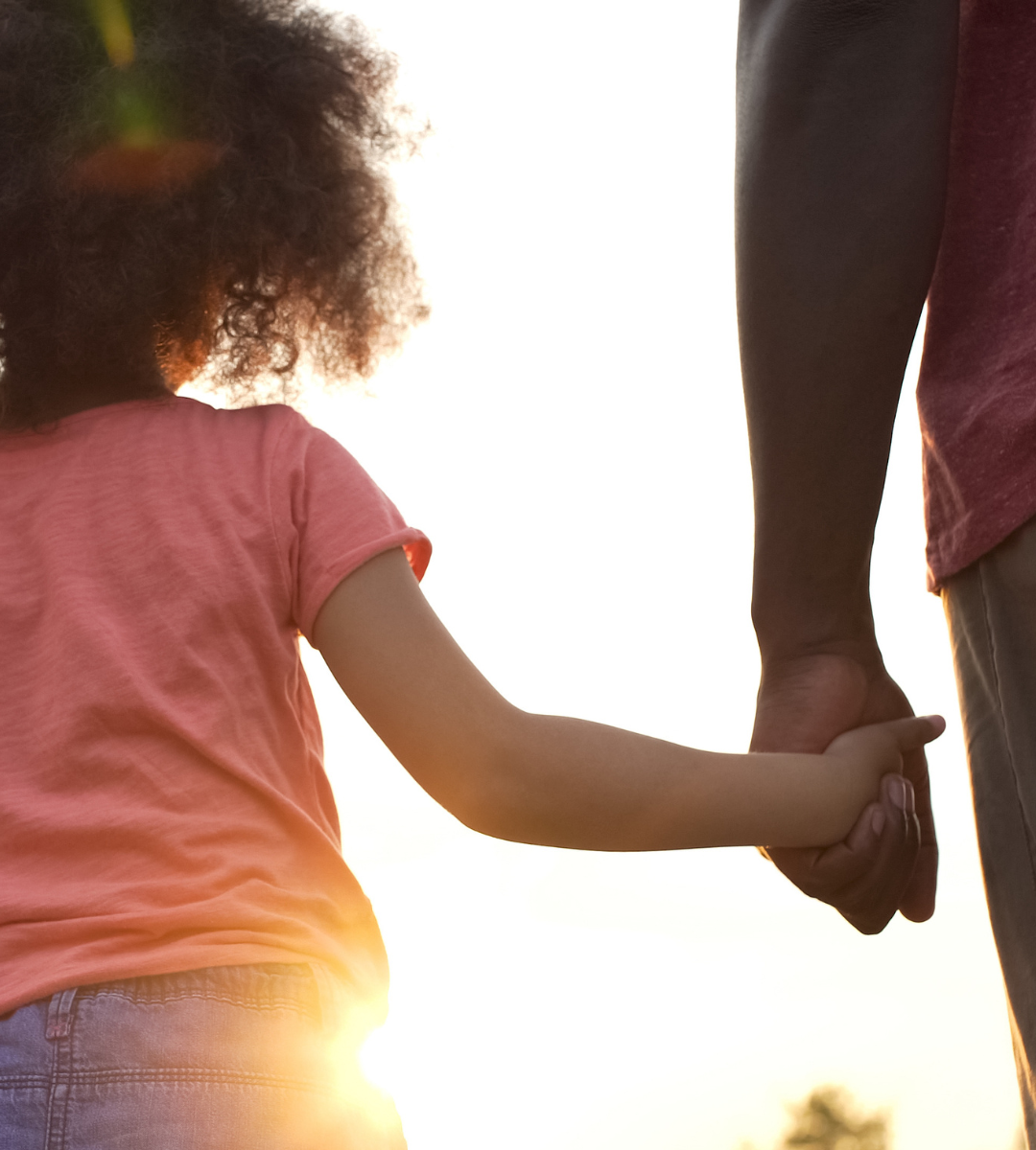 A young girl holding an adult's hand during sunset, with the sun in the background.
