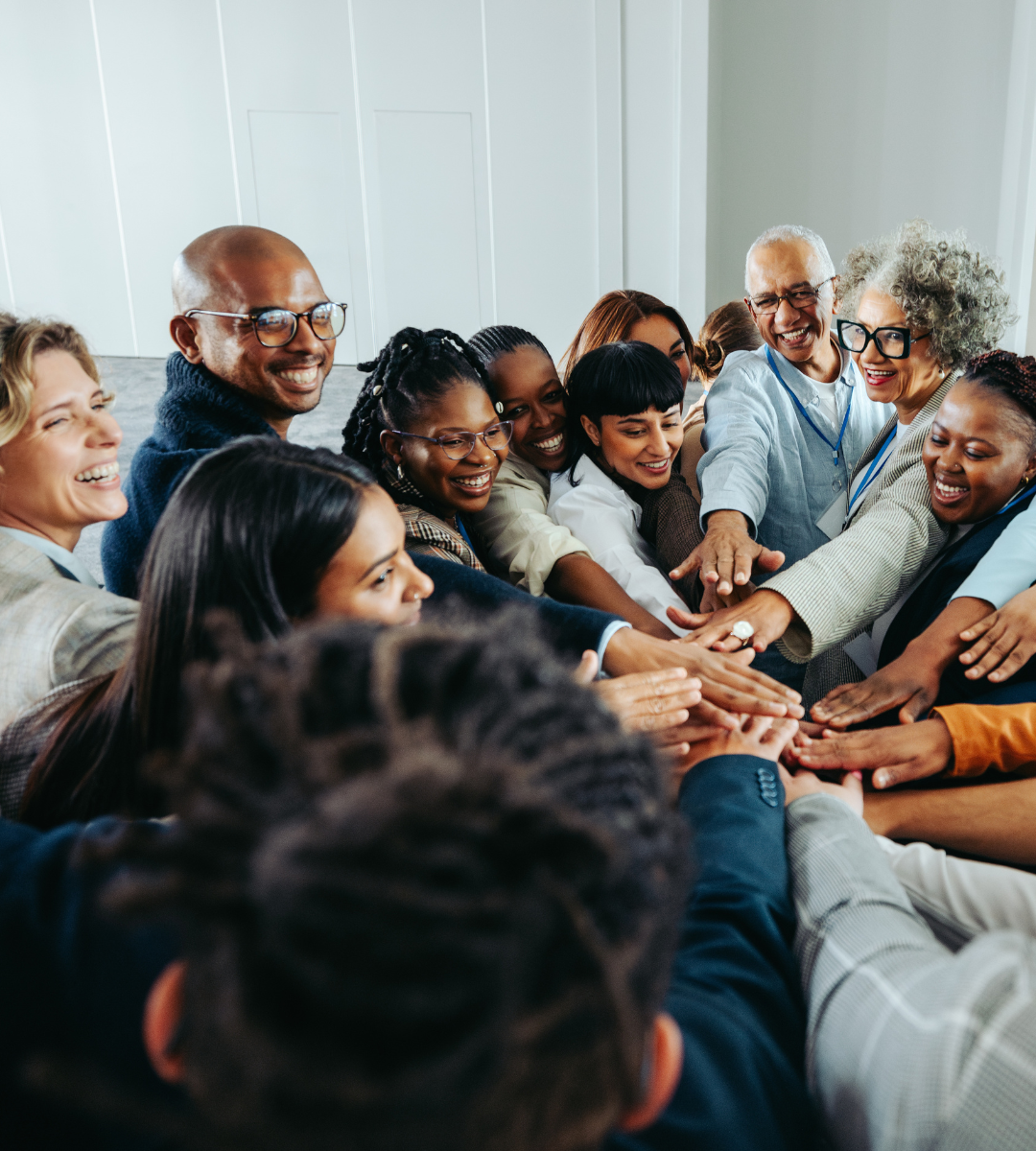 Group of diverse smiling people with hands stacked in the center, celebrating teamwork in an indoor setting.