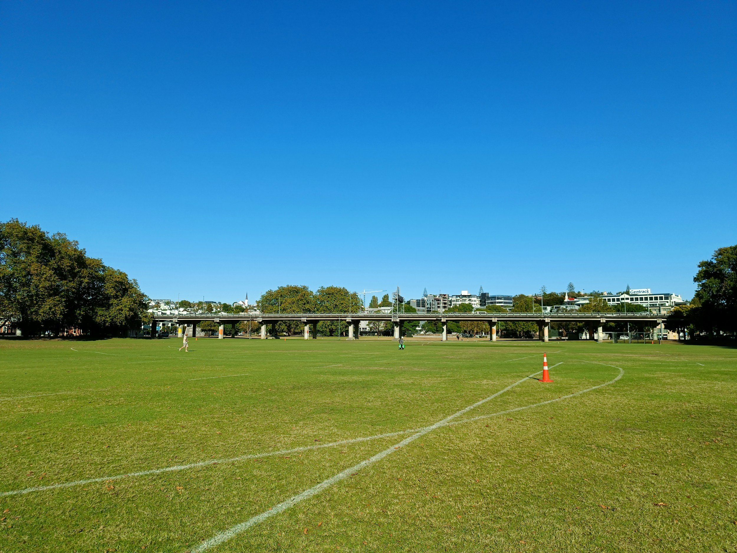 A green field with white painted lines, orange cones, and a few small people. In the background, there are trees, a bridge, and buildings under a clear blue sky.
