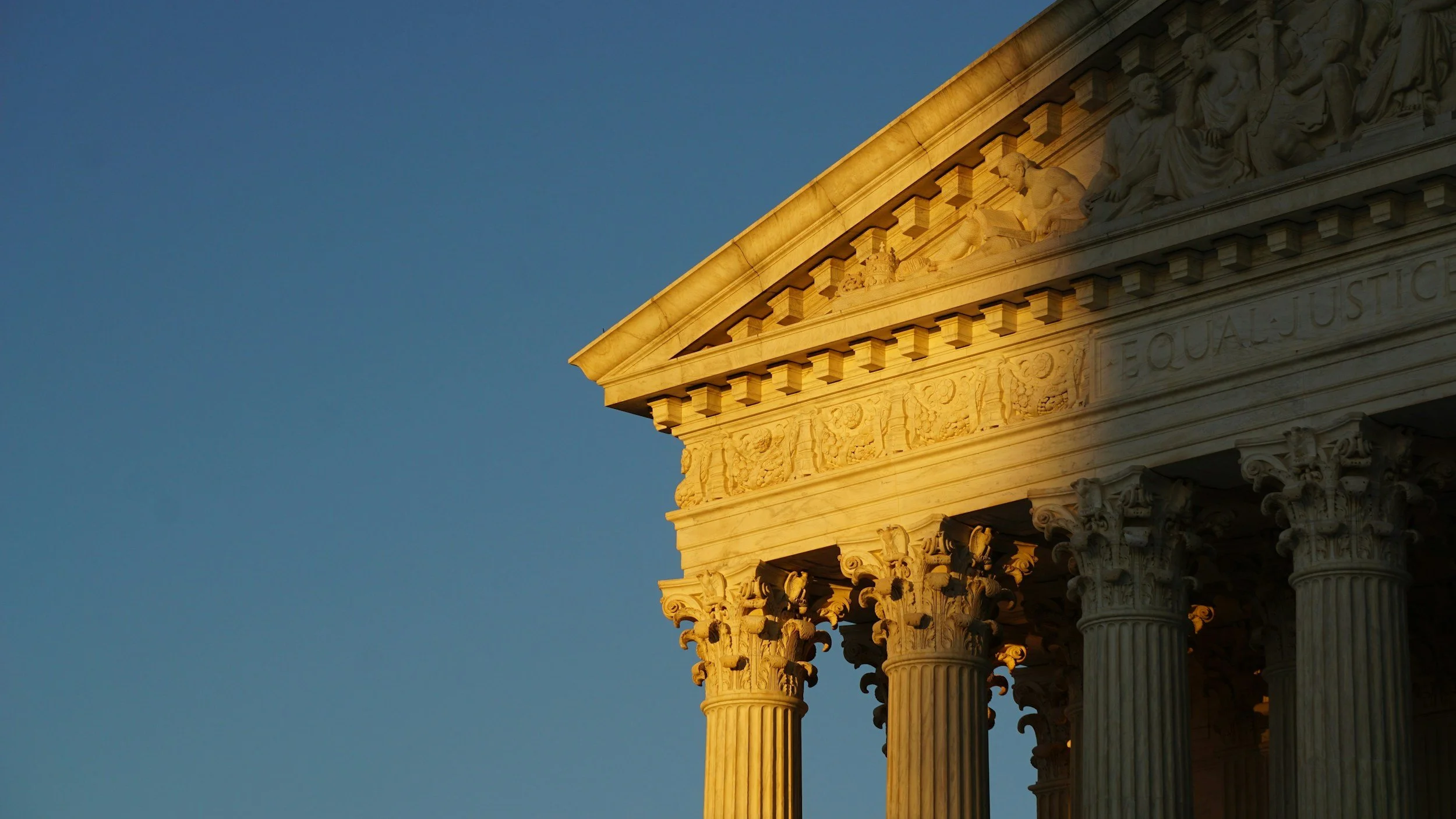 Close-up of the upper part of a historic building with columns and detailed sculptures, illuminated by sunlight against a clear blue sky.