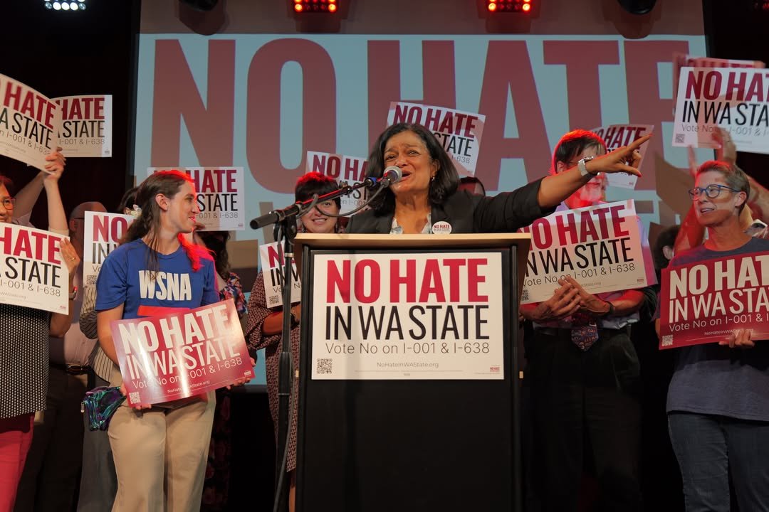 A woman speaking at a podium with a microphone, surrounded by people holding signs that read 'No Hate In Wastate.' The background has a large display with similar message. The scene appears to be at a rally or political event advocating against hate and supporting voting on initiatives I-001 and I-638.