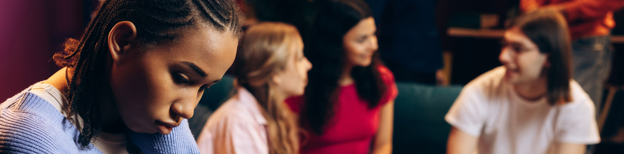 A young woman with braided hair looking down, sitting at a table with four other women, all appearing to be in a casual or social setting.