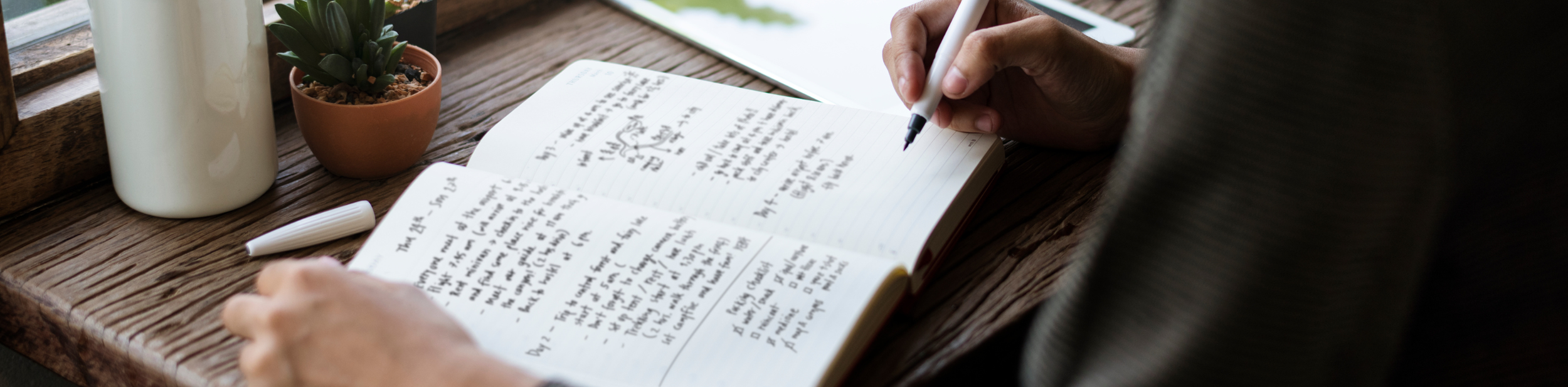 A person writing in a notebook with a black pen on a wooden table. There are two potted plants, a white bottle, and a white marker on the table.
