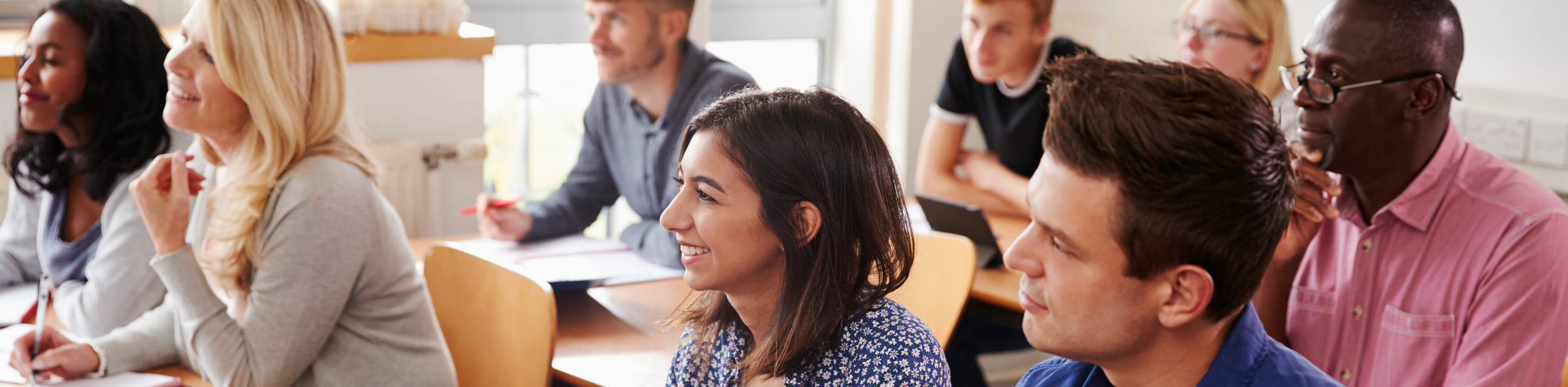A diverse group of adults attending a classroom seminar, sitting at tables and listening attentively.