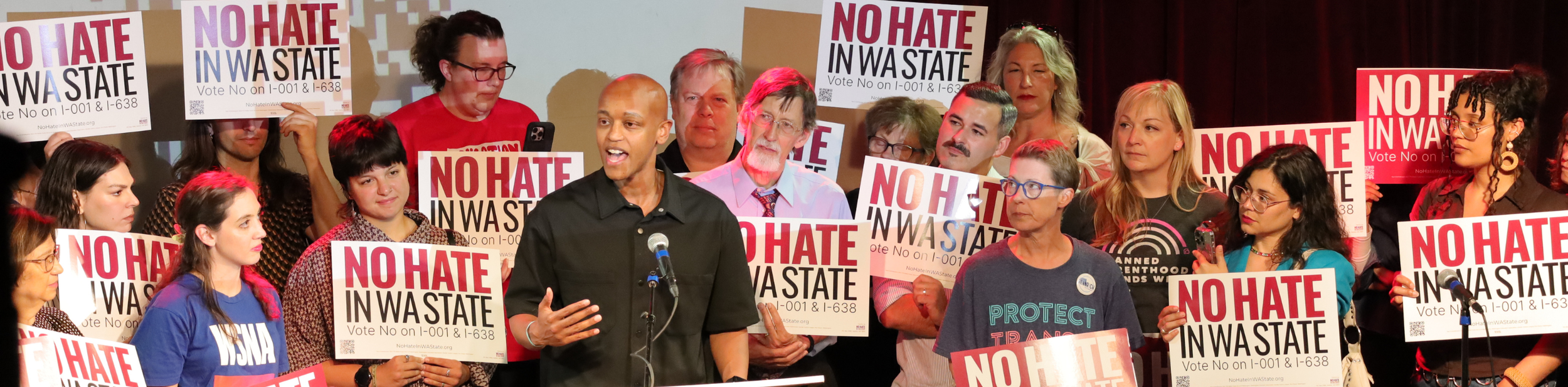 A diverse group of people at a rally or protest holding signs that read 'NO HATE IN WASTE' with a man speaking at a microphone in the foreground.