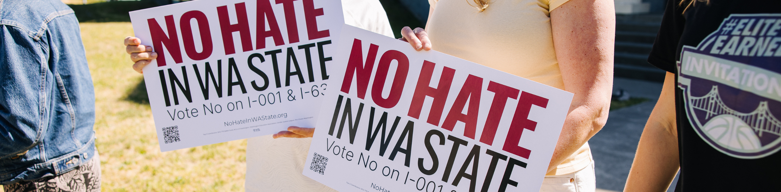 Two people holding signs that say "NO HATE IN WASHINGTON" with a subtitle "Vote No on I-001 & 1-63" during a protest or rally. The background shows grass and a person wearing a denim jacket and another wearing a shirt with a basketball emblem.