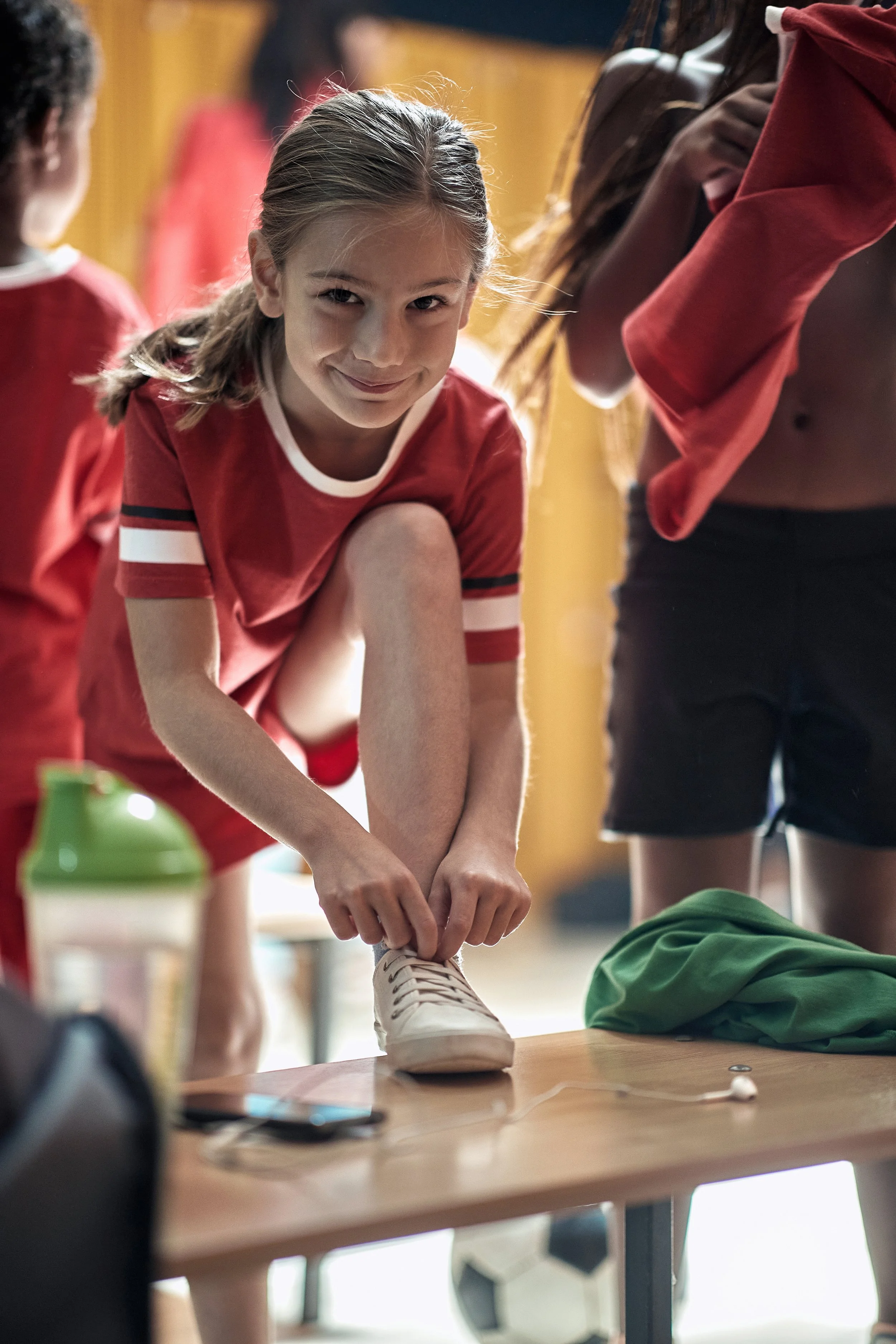 A young girl in a red sports jersey is crouching on a table, tying her white sneaker. Other children are visible in the background, and the setting appears to be a sports or gym environment.
