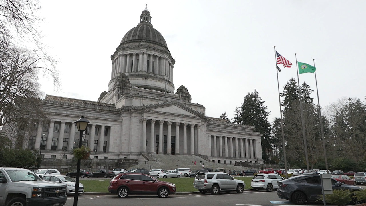The State Capitol building with a large dome and columns, flags flying outside, parked cars in front, and trees surrounding the area.