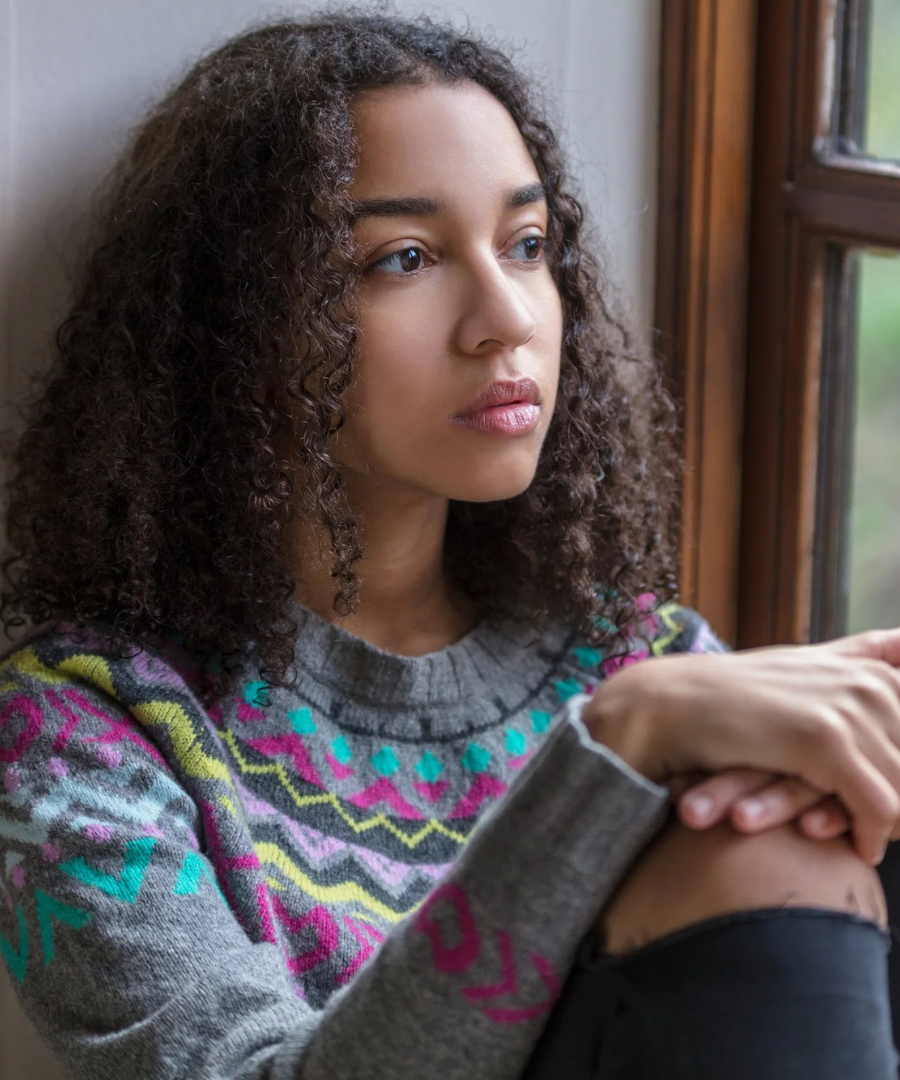 A young woman with curly hair looking out a window, wearing a colorful patterned sweater.