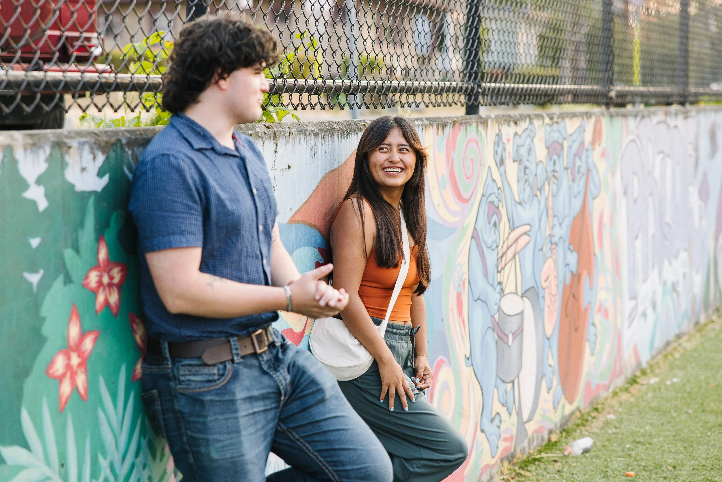 Two young people, a man with curly dark hair wearing a blue shirt and a woman with long dark hair in an orange top, sitting and leaning against a colorful mural on a concrete wall near a chain-link fence, outdoors in daytime.