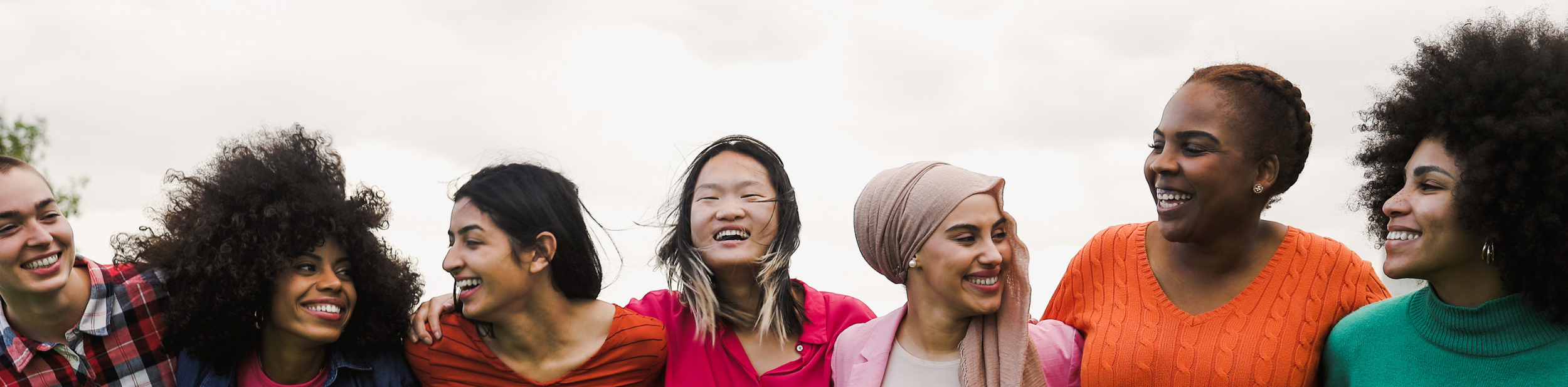 Group of diverse women smiling and laughing outdoors on cloudy day.