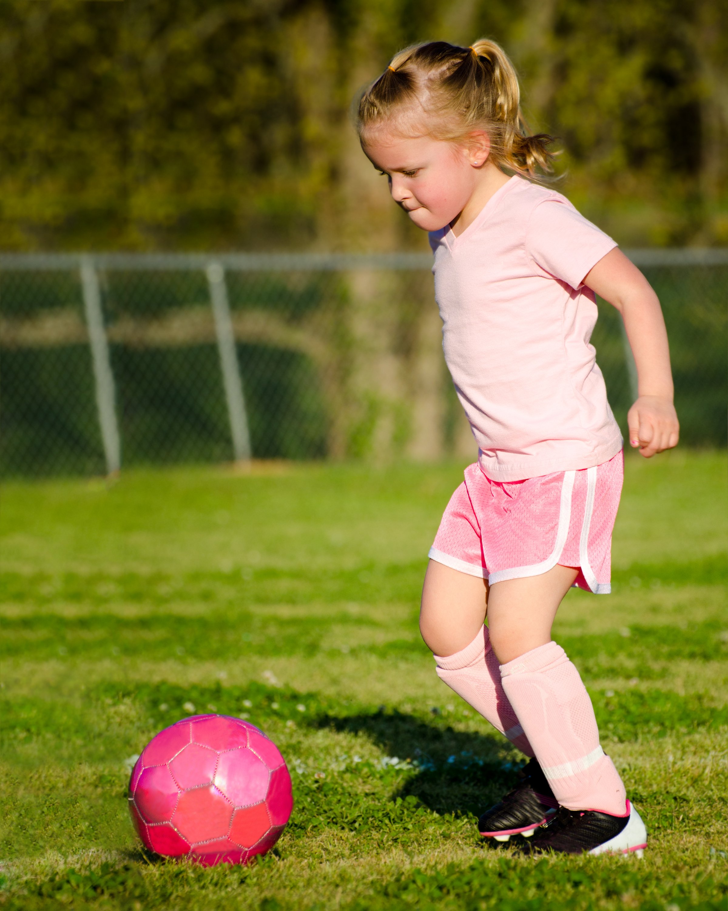 Young girl in pink sportswear playing soccer with a pink ball on a grassy field.