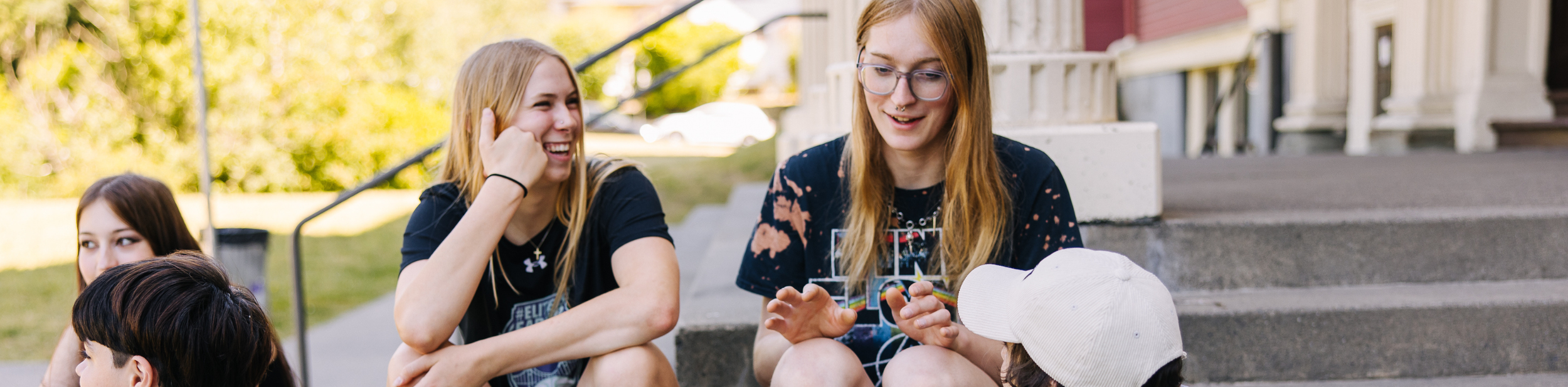 Four young women and a young boy sitting on stairs outside, engaging in conversation and smiling, with trees and houses in the background.