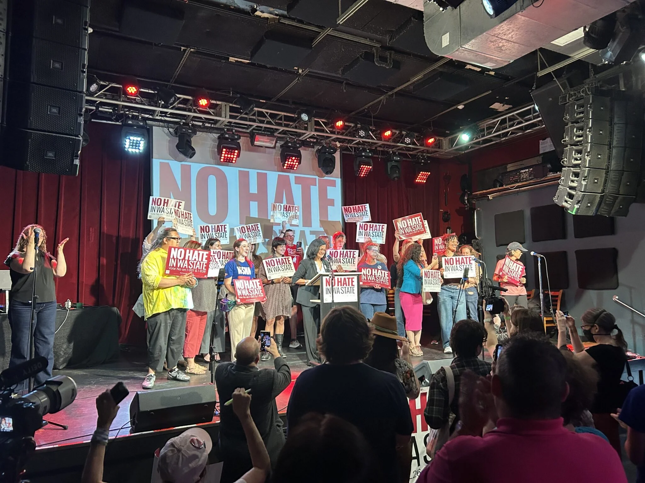 A group of people on stage protesting with signs that say 'NO HATE IN WASTATE.' The stage has a large sign that reads 'NO HATE' and is equipped with professional lighting and audio equipment. The audience is visible in front of the stage.
