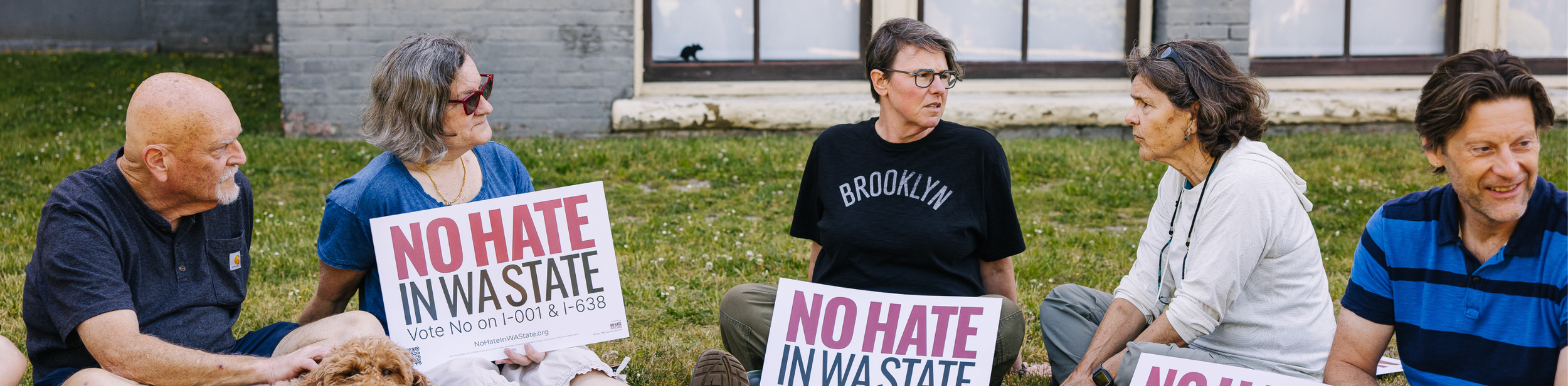 Group of people sitting on grass holding signs that read 'NO HATE IN Washington,' during a protest or rally.