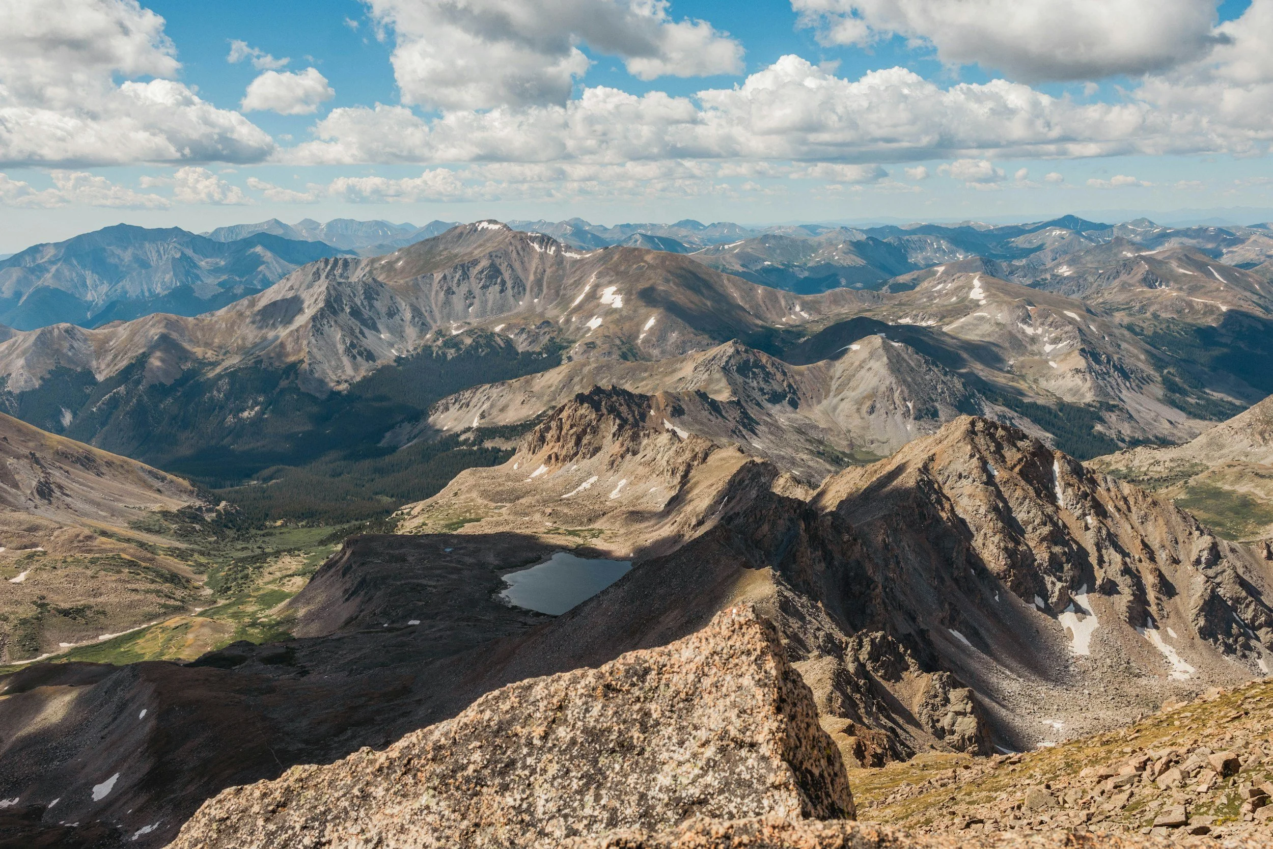 A panoramic view of rugged mountain peaks with patches of snow, a small mountain lake in the foreground, and a partly cloudy sky overhead.