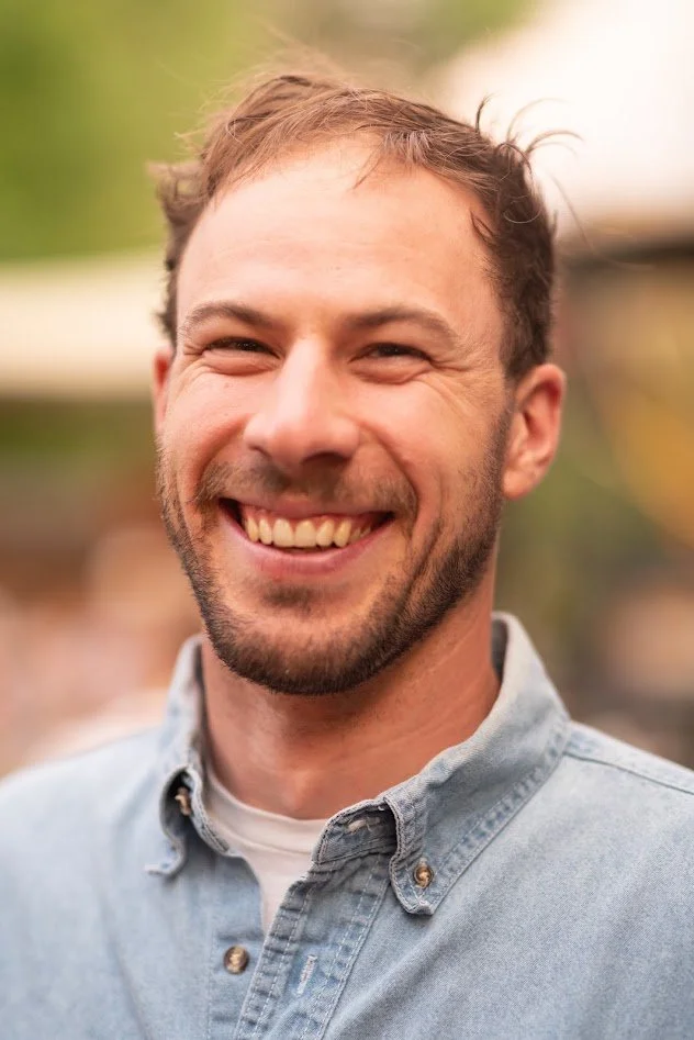 Smiling man with short brown hair and a beard, wearing a denim shirt in an outdoor setting.