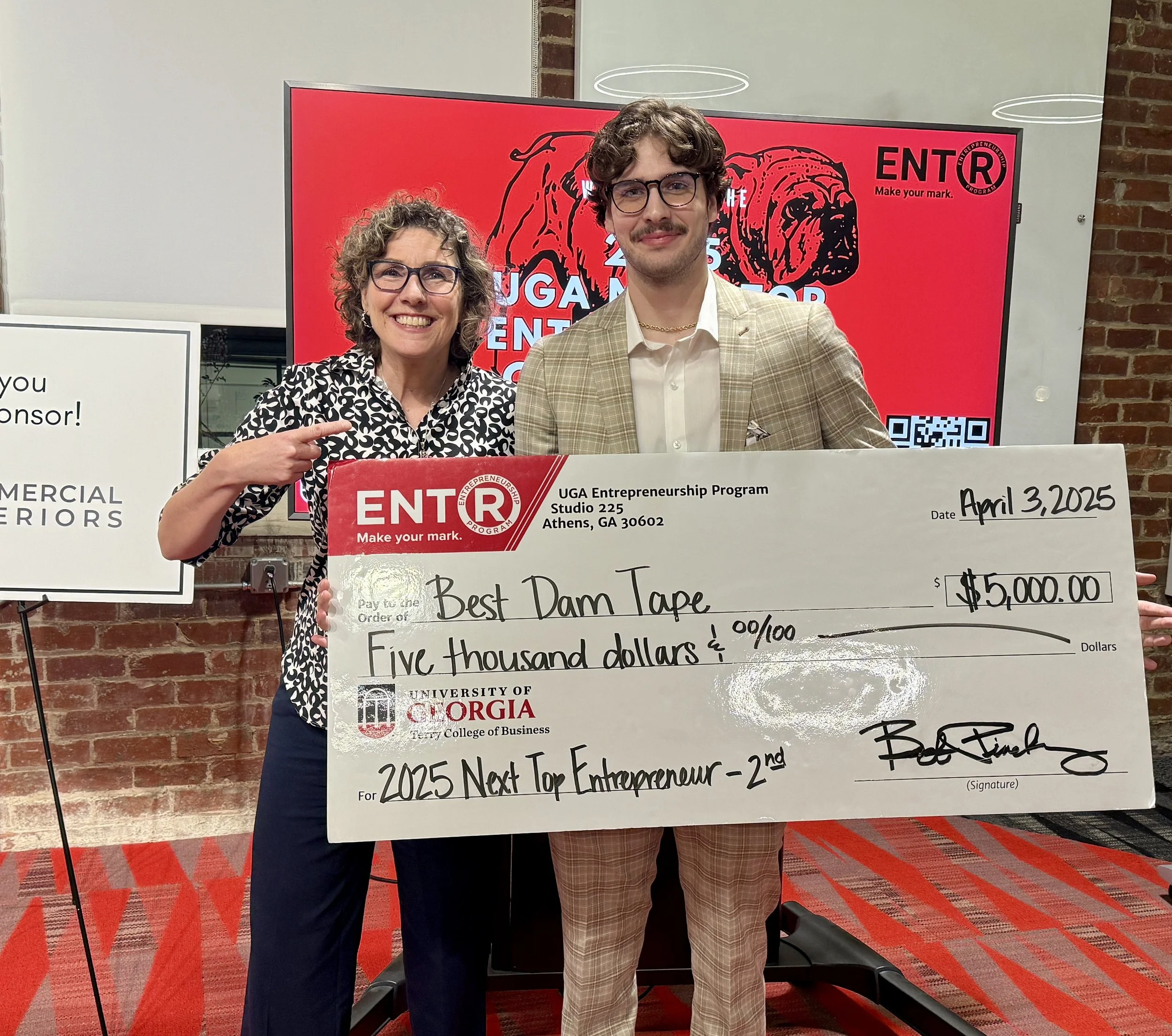 Two people, a woman with curly hair and a man with glasses, smiling and holding a large check for five thousand dollars at an event. The background features a red sign with black, white, and red text and graphics, including a bulldog illustration, and a brick wall. The woman is pointing at the check, which is made out to the recipient for the 2025 Next Top Entrepreneur competition.