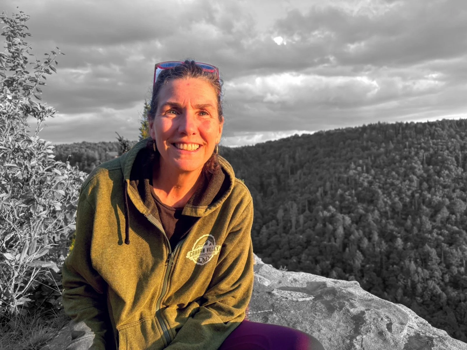 A woman smiling and sitting on a rock with a mountainous landscape in the background. The sky is cloudy, and the photo is partly in black and white with the woman in color.