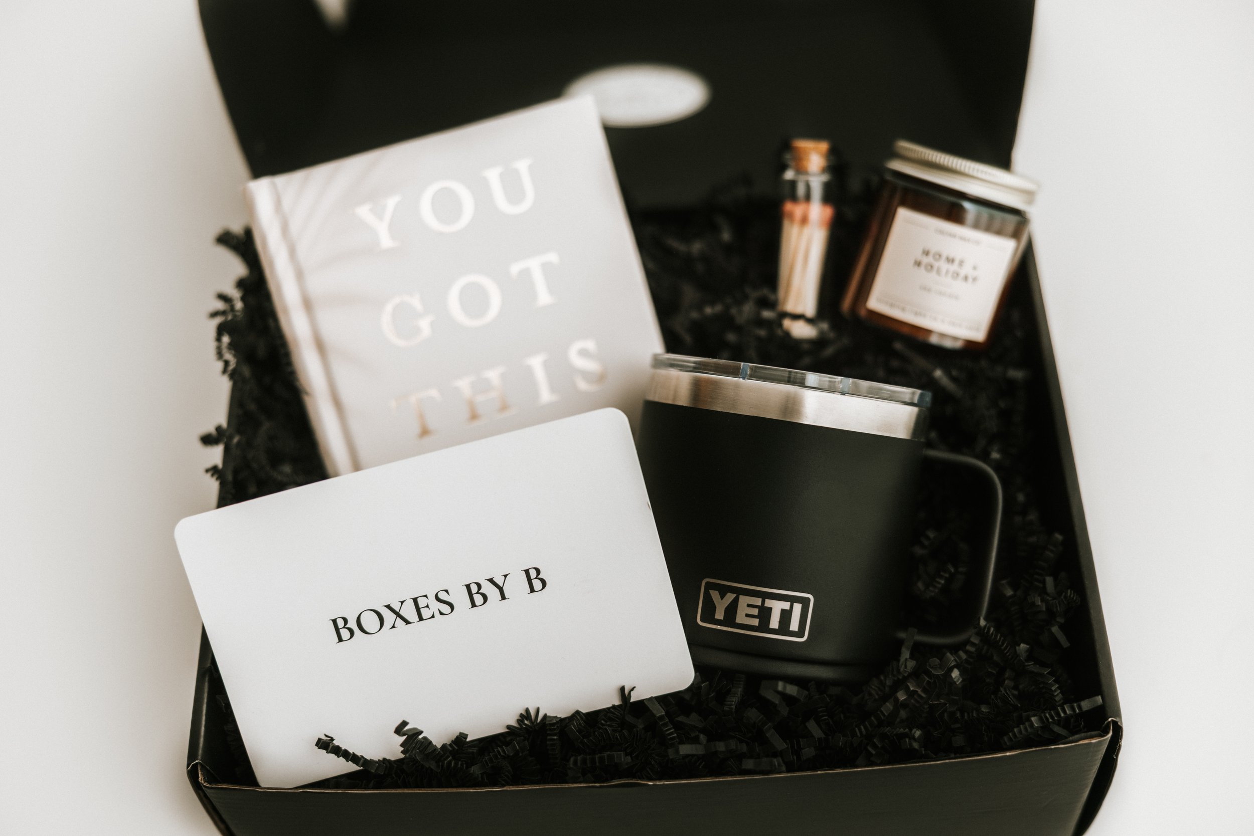 A black gift box with crinkle paper fill contains a white card that reads 'BOXES BY B', a black YETI mug, a white candle labeled 'HOME FOR THE HOLIDAYS', a small jar, and a small container of matches. Tucked against the side is a book with the words 'YOU GOT THIS' on the cover.