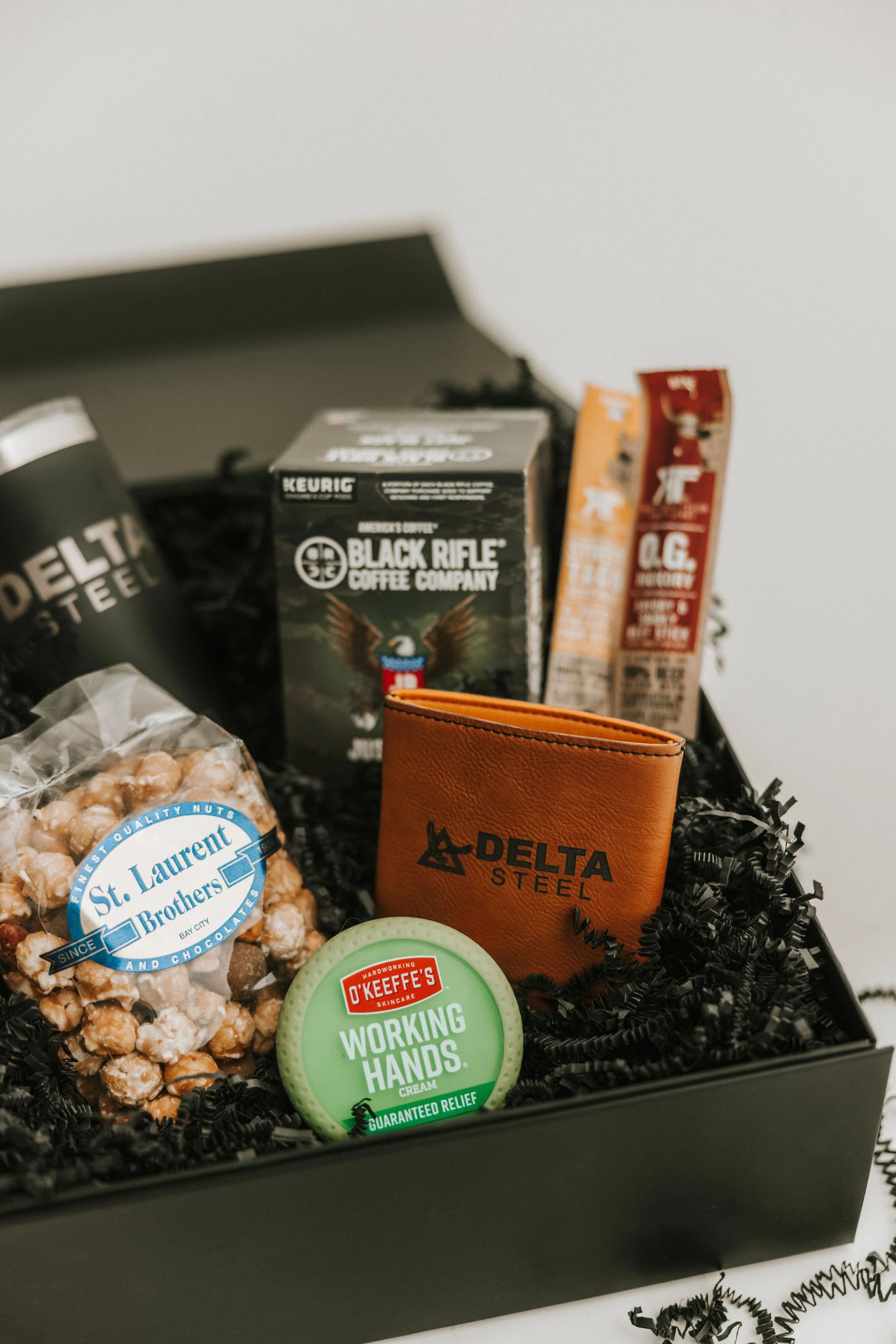 Gift box containing coffee, snacks, a leather wallet with 'Delta Steel' logo, and hand cream on black shredded paper.