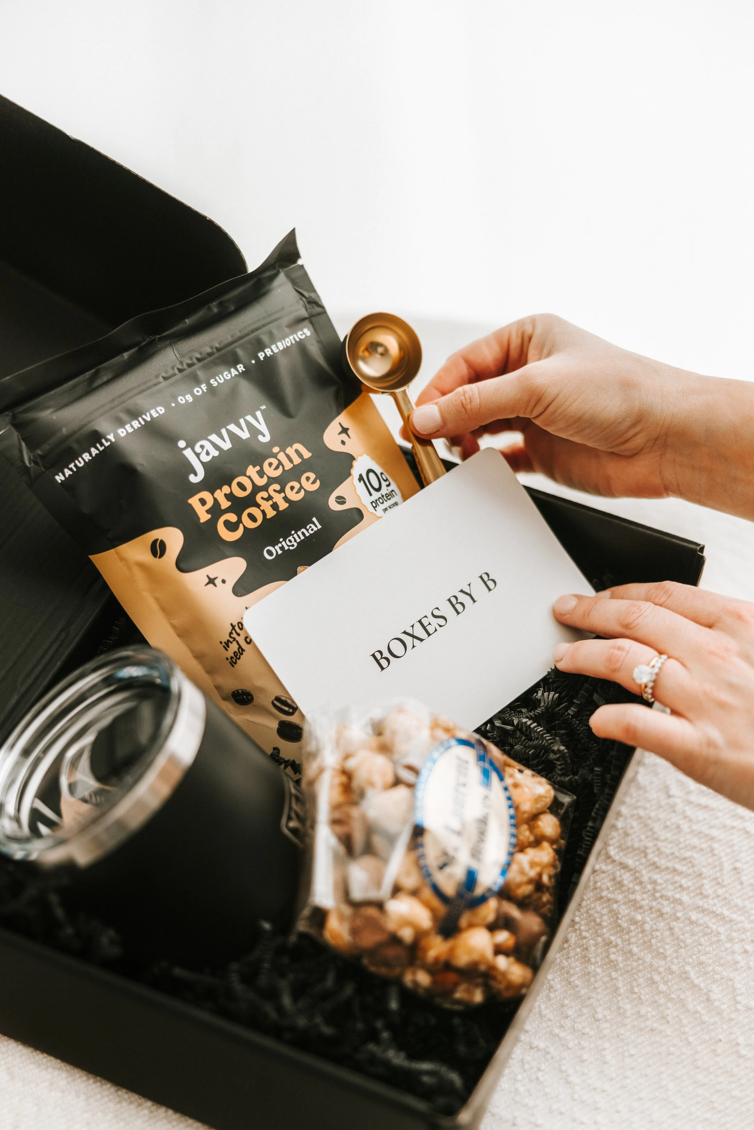 A gift box labeled 'BOXES BY B' contains a bag of Javvy protein coffee, a small jar, and a box of popcorn, with a hand holding a gold spoon.