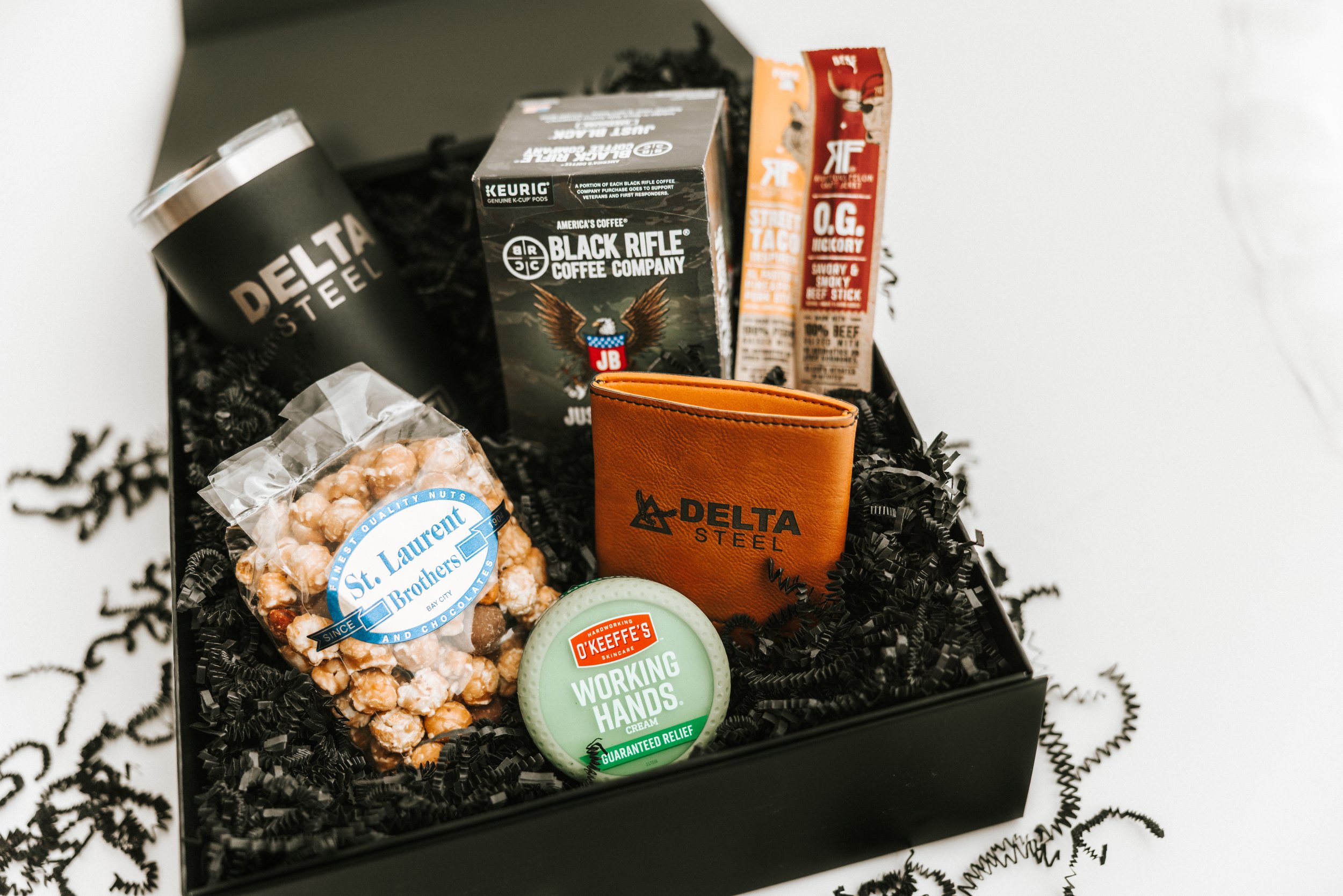 Gift box containing a Delta Steel wallet, a tin of O'Keeffe's Working Hands cream, a bag of St. Laurent Nuts, a Delta Steel branded travel tumbler, a box of Black Rifle Coffee, and two cougar flavored snack sticks.