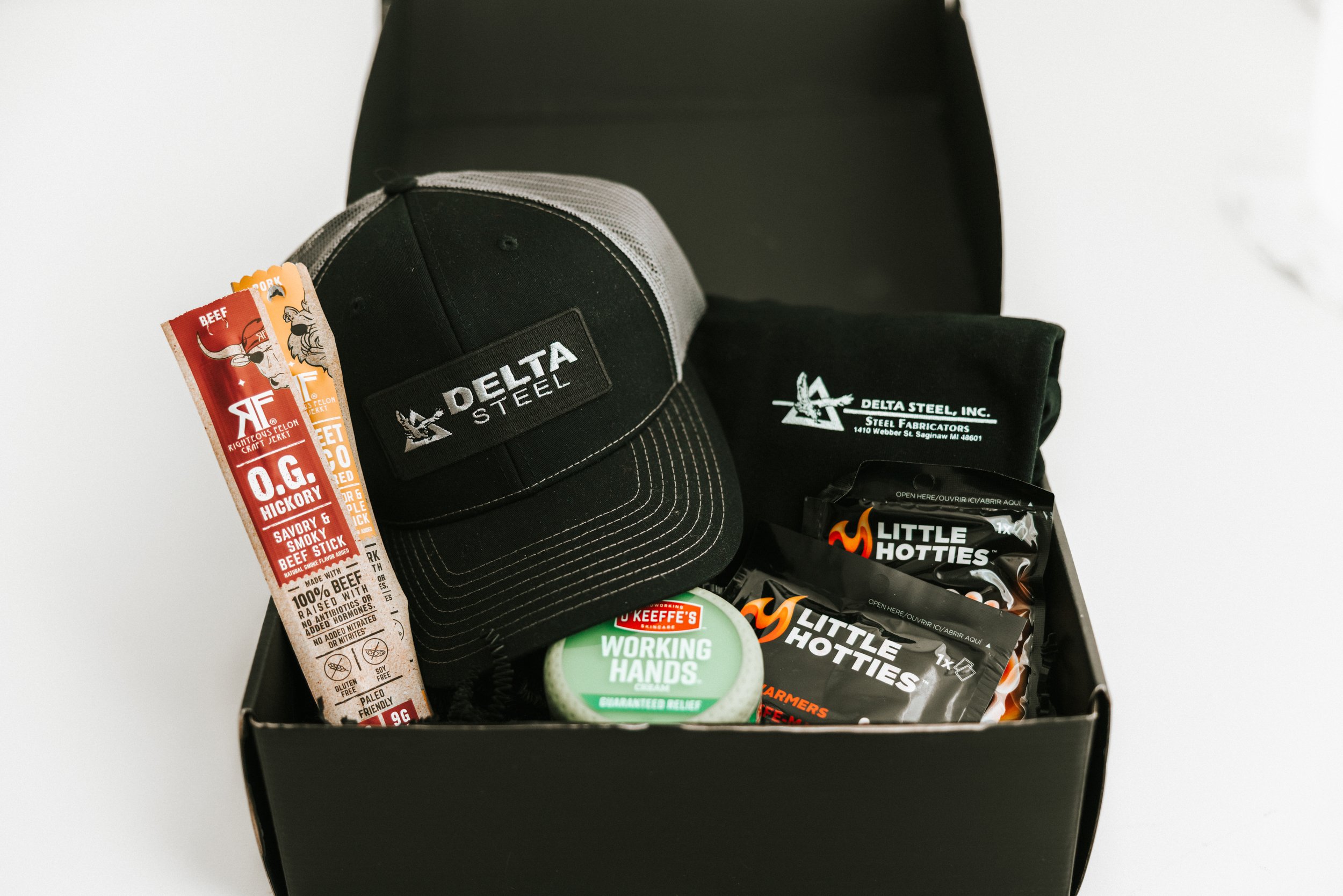Open black box containing a gray and black Delta Steel cap, a black Delta Steel cloth, a pack of Little Hotties hand warmers, a Keeffe's Working Hands cream, a bag of beef jerky, and a pack of beef jerky in orange and black packaging.