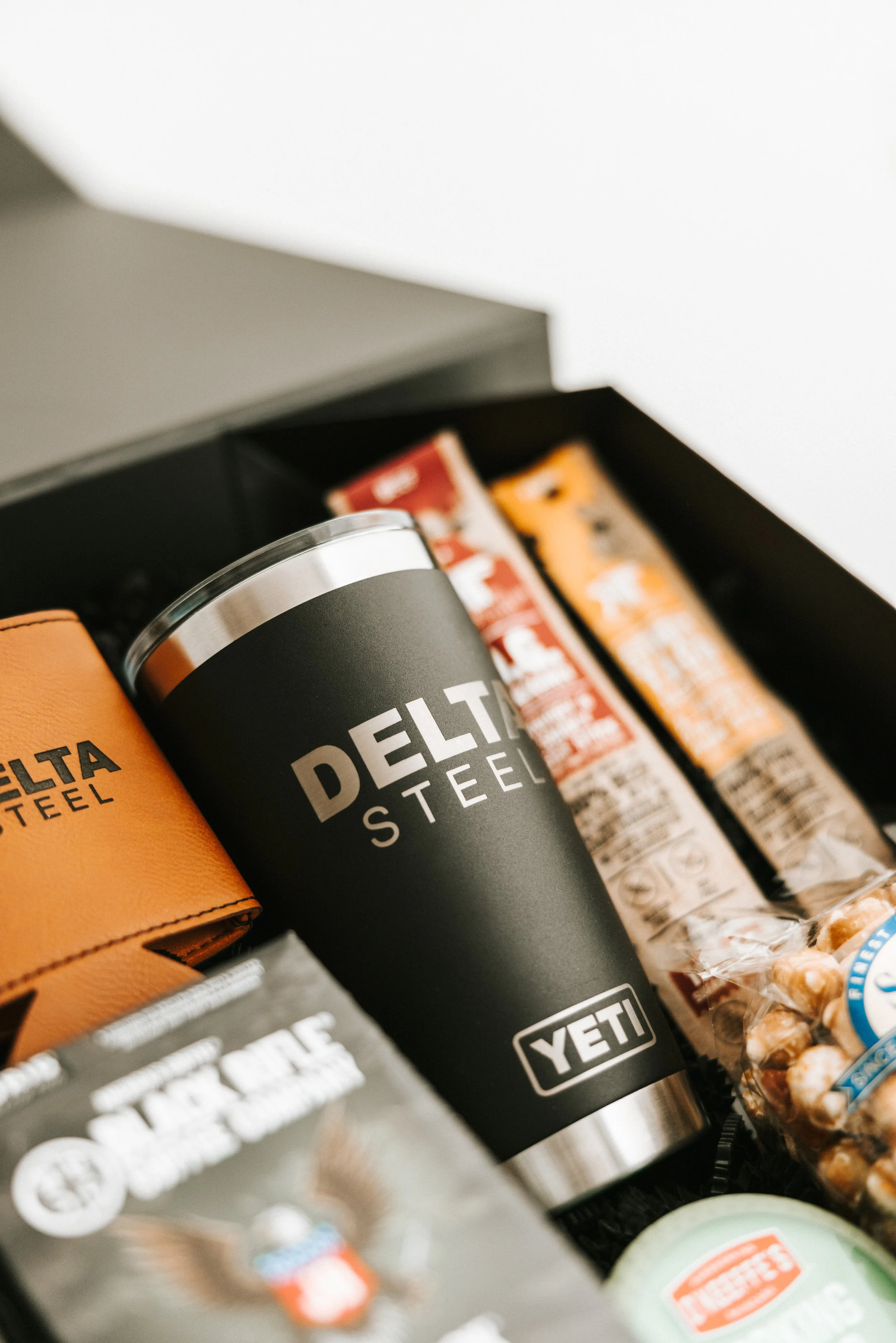 A close-up of a black YETI tumbler with 'Delta Steel' branding, surrounded by snacks, energy bars, and a tan wallet inside a box.