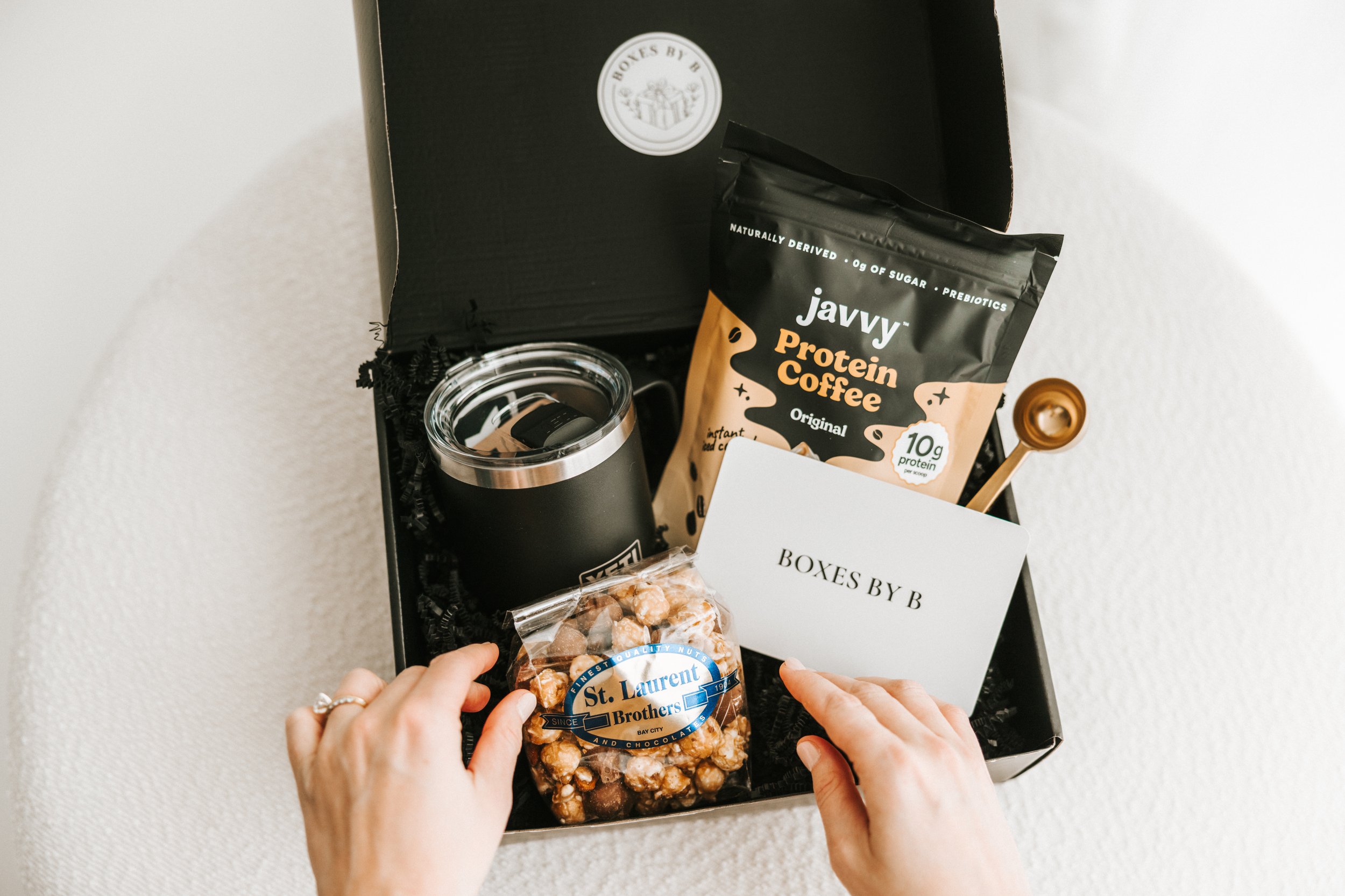 Open black box with assorted items including a jar, a bag of protein coffee, a bag of mixed nuts, a white note card with 'BOXES BY B' written on it, and a small copper-colored scoop, all arranged on a white surface.