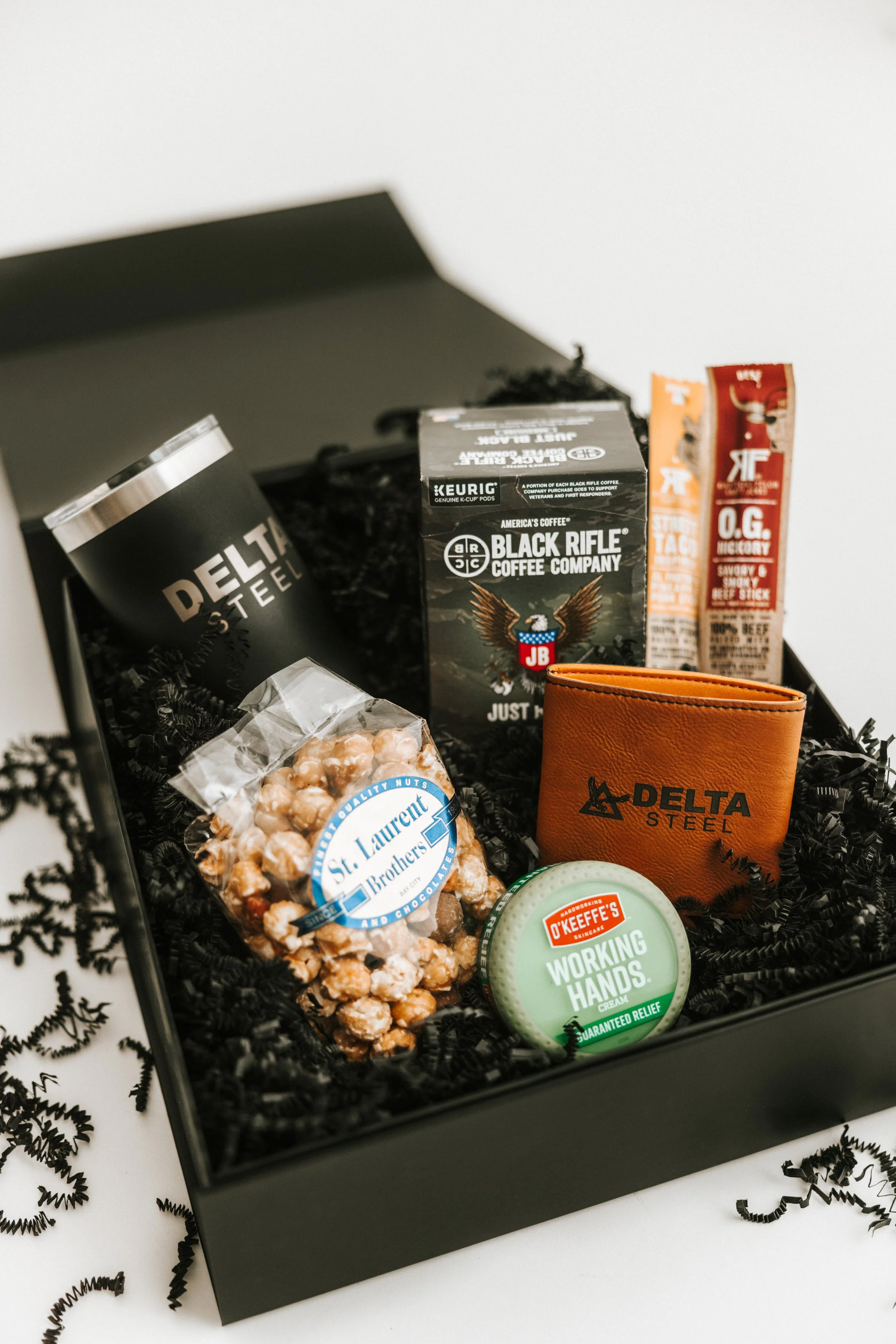 A gift box containing various products including a coffee tin, a box of Black Rifle Coffee, a container of hand cream, a leather wallet with Delta Steel logo, a bag of caramel popcorn, and energy bars, all arranged on shredded black paper.