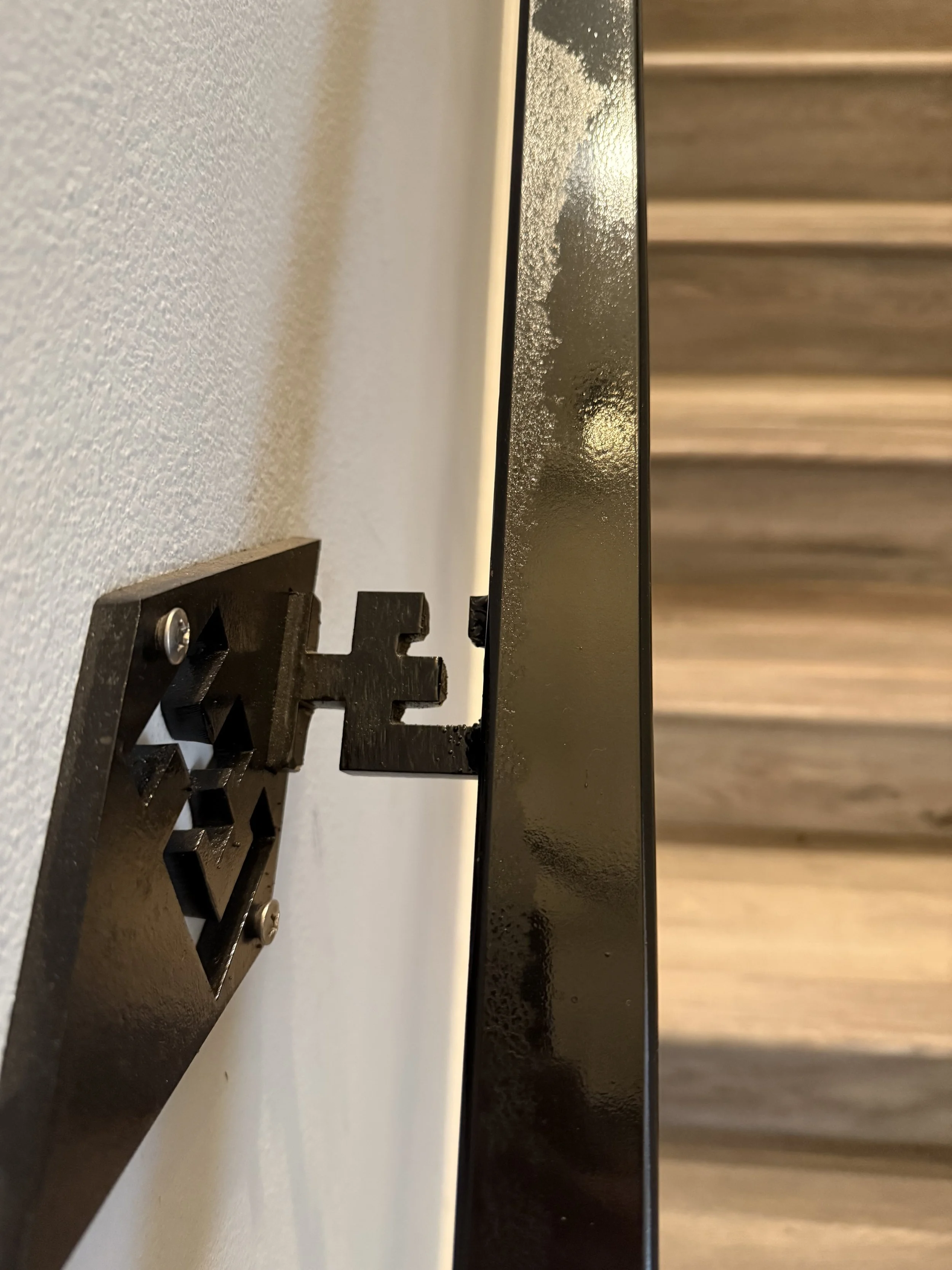 Close-up of a black wall-mounting bracket attached to a wall with an electrical outlet, with a black metal object partially visible next to the wall.