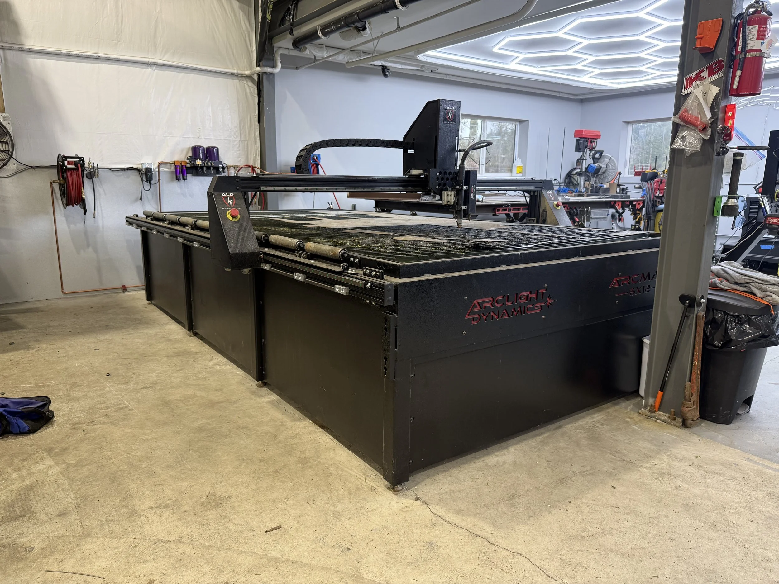 A large black laser cutting machine labeled 'ArcLight Dynamics' in a workshop with tools, fire extinguisher, and other equipment.