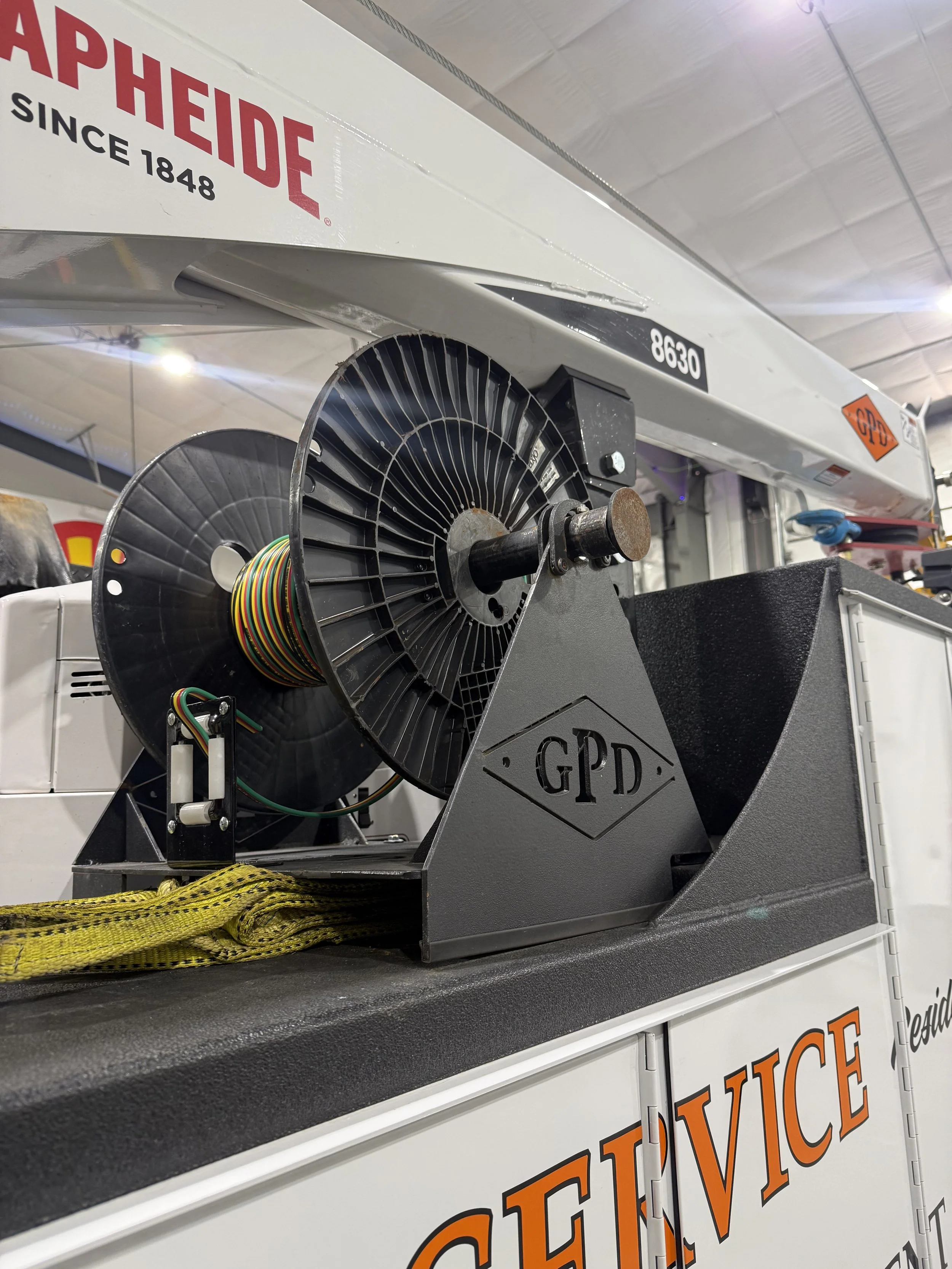 Close-up of industrial equipment with wiring and a reel mounted on a black and gray stand with GPD logo in an indoor setting with a large white overhead structure.